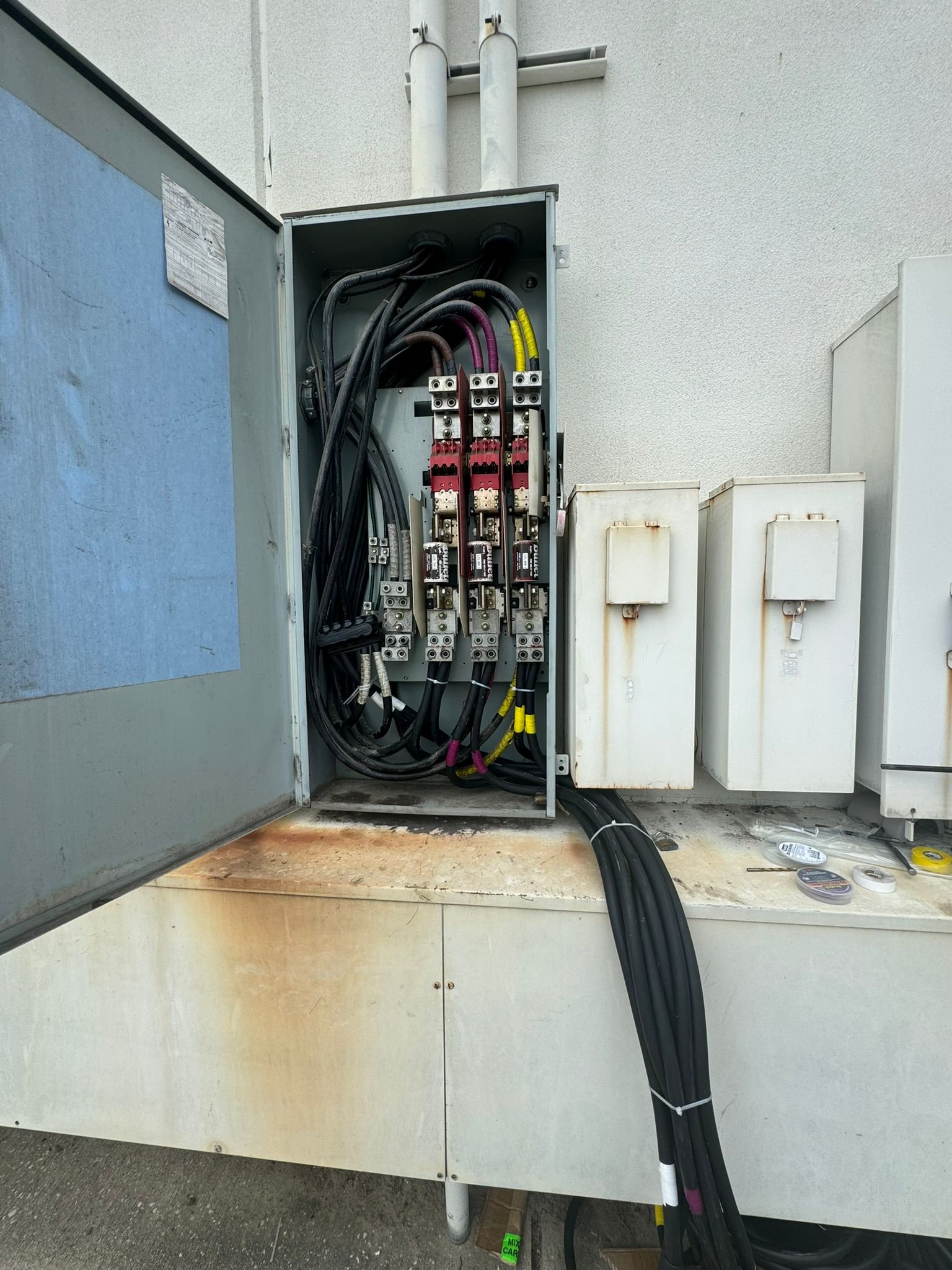 An electrical box containing black and colored wires, some with yellow protective coverings, inside a metal cabinet mounted on a wall with a concrete surface below.