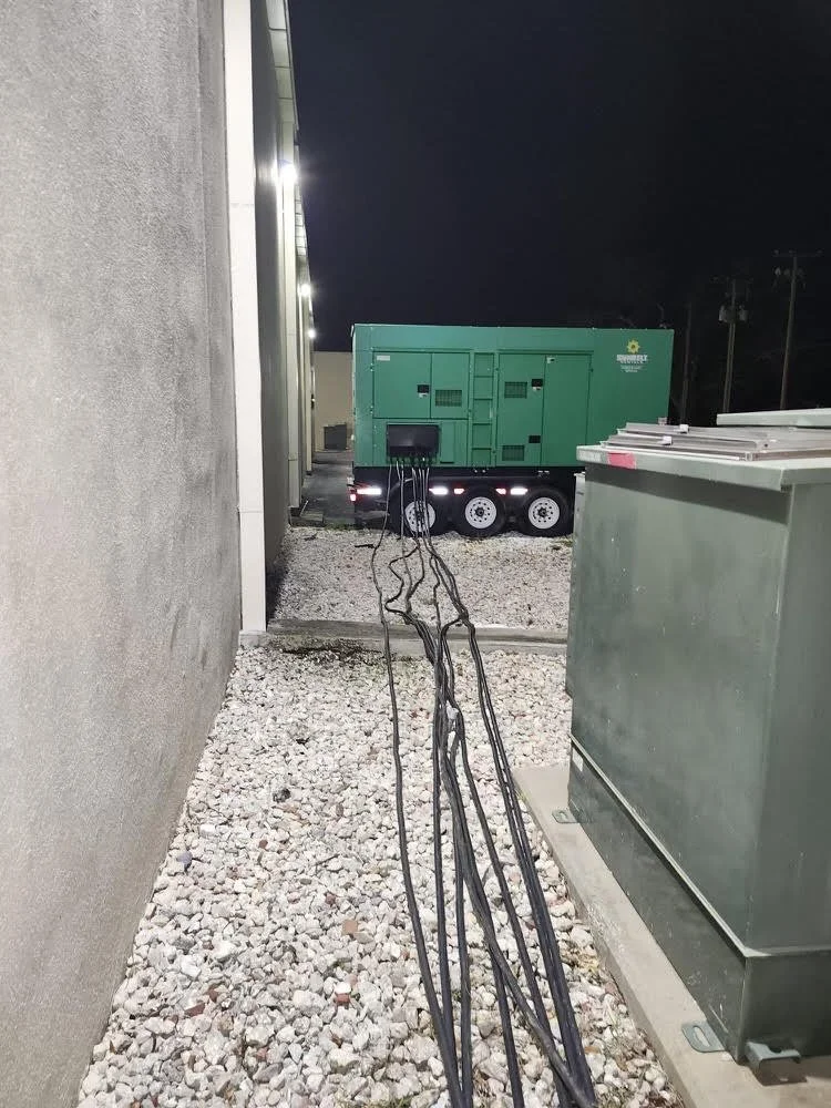 Nighttime scene with multiple black cables hanging down between a gray building wall and a gray utility box, leading to a large green generator on a trailer in a gravel-covered area.