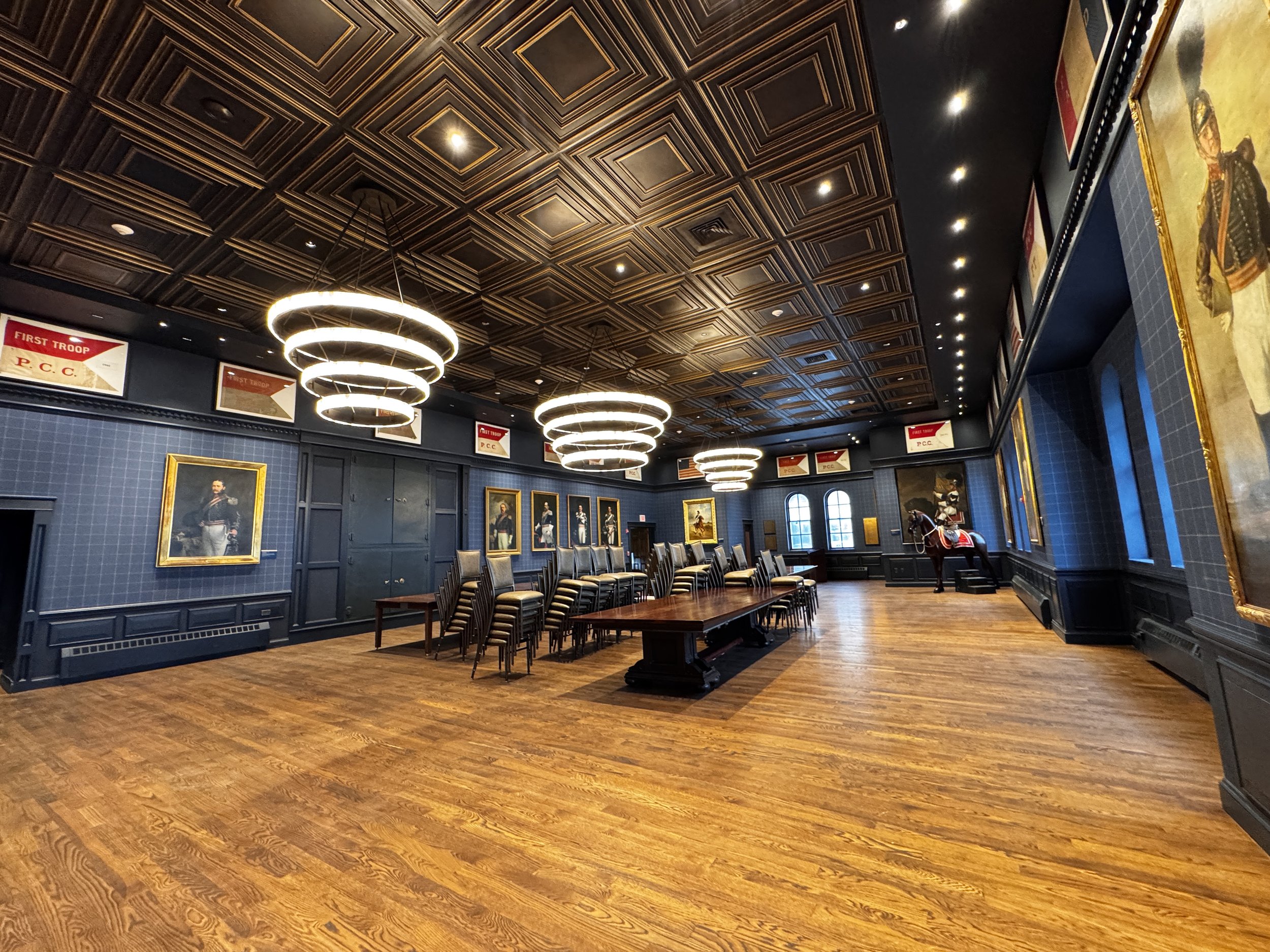 Interior of a room with dark blue walls, wooden flooring, paintings in golden frames, modern circular chandeliers, and a row of chairs around a large wooden table.