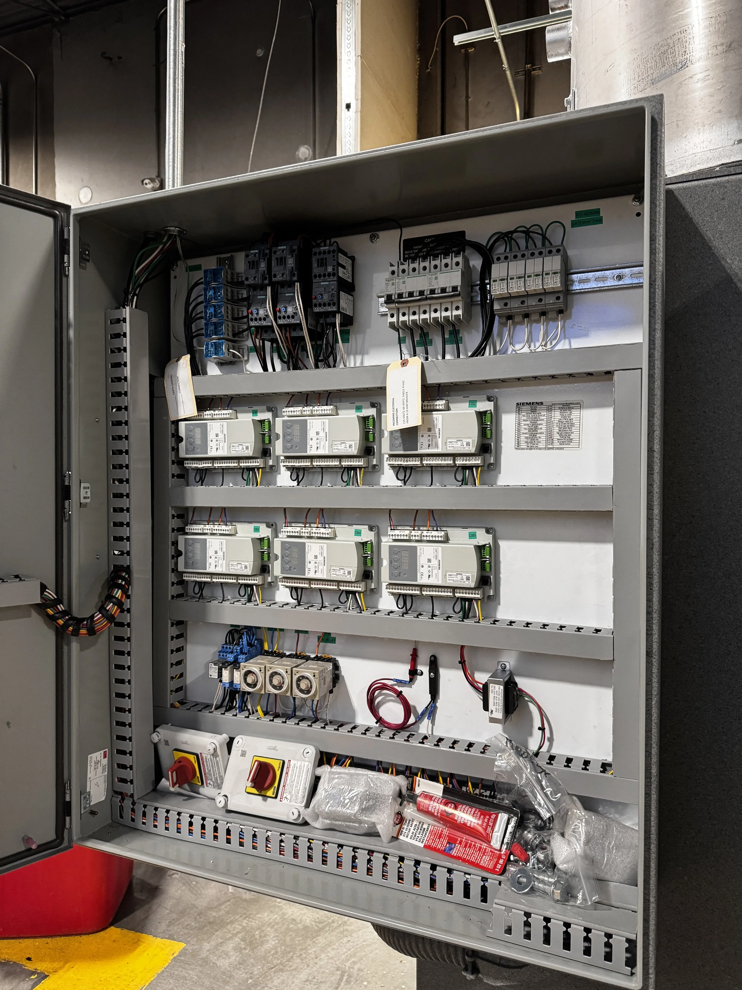 An open electrical control panel with various wires, circuit breakers, relays, and electrical components inside. Fire extinguisher and tools are nearby.