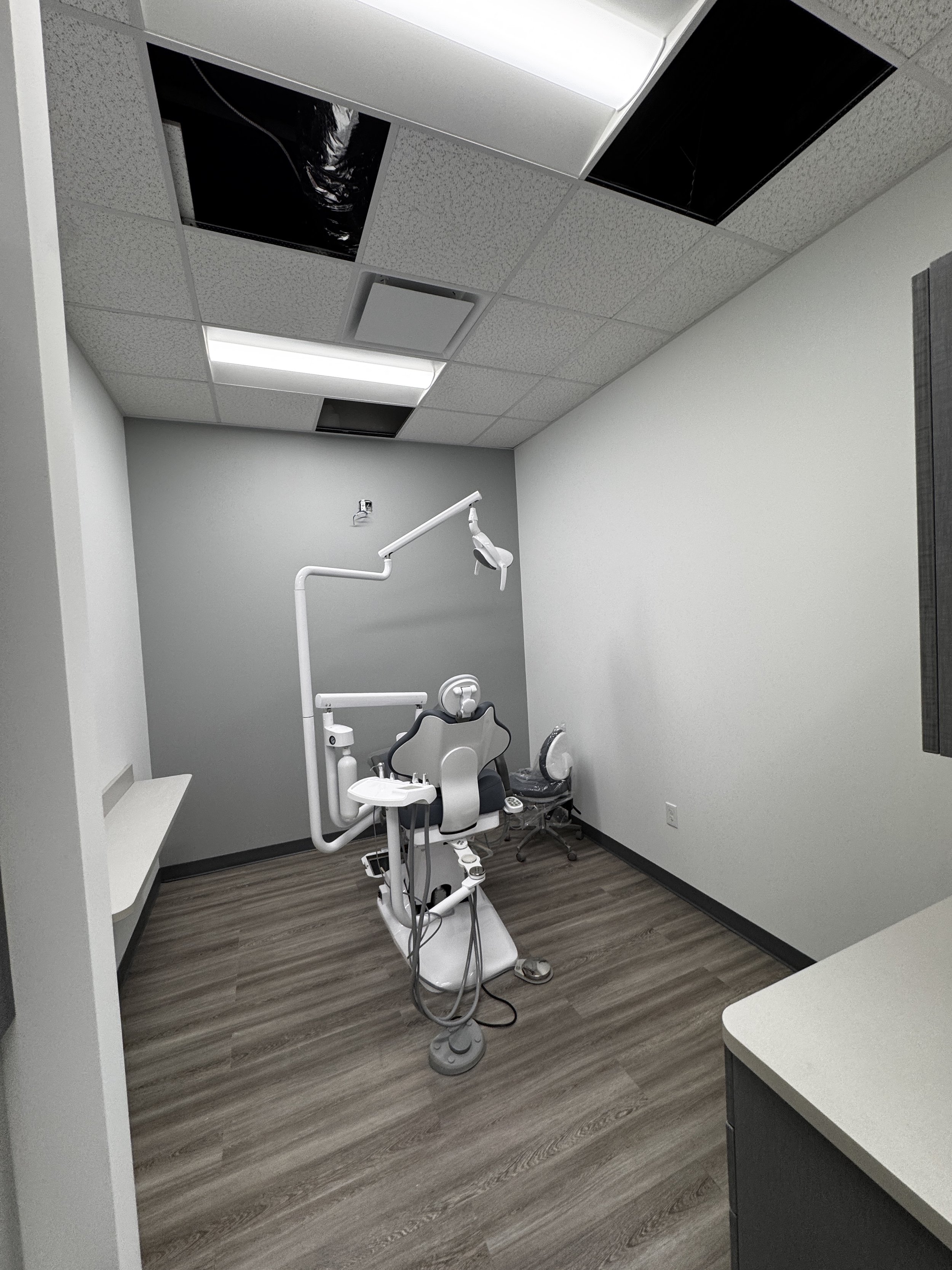 A dental examination room with white walls, a dental chair, and various dental equipment. The ceiling has some ceiling tiles missing, revealing ductwork and lighting fixtures.