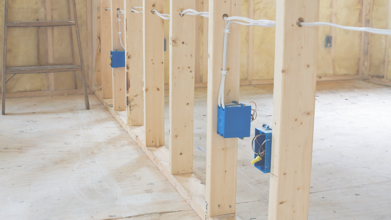 Interior view of a building under construction with exposed wooden studs and electrical wiring, with a step ladder on the left side.