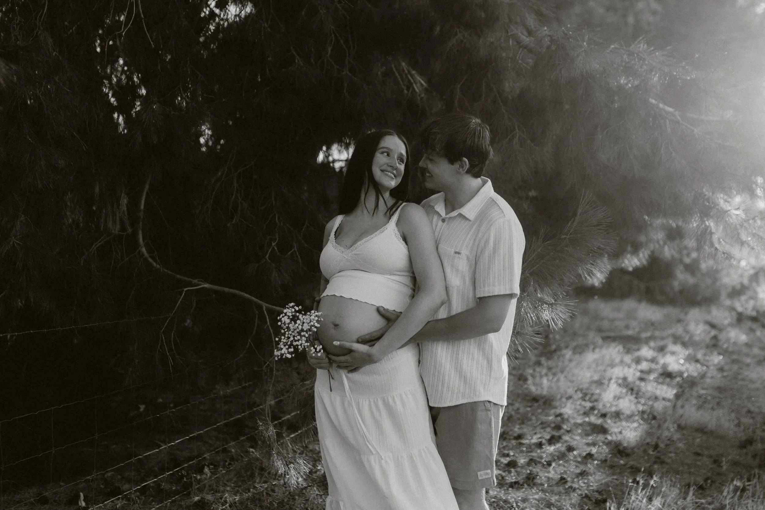 A couple standing close together outdoors, with the woman showing a pregnant belly, surrounded by trees and foliage.