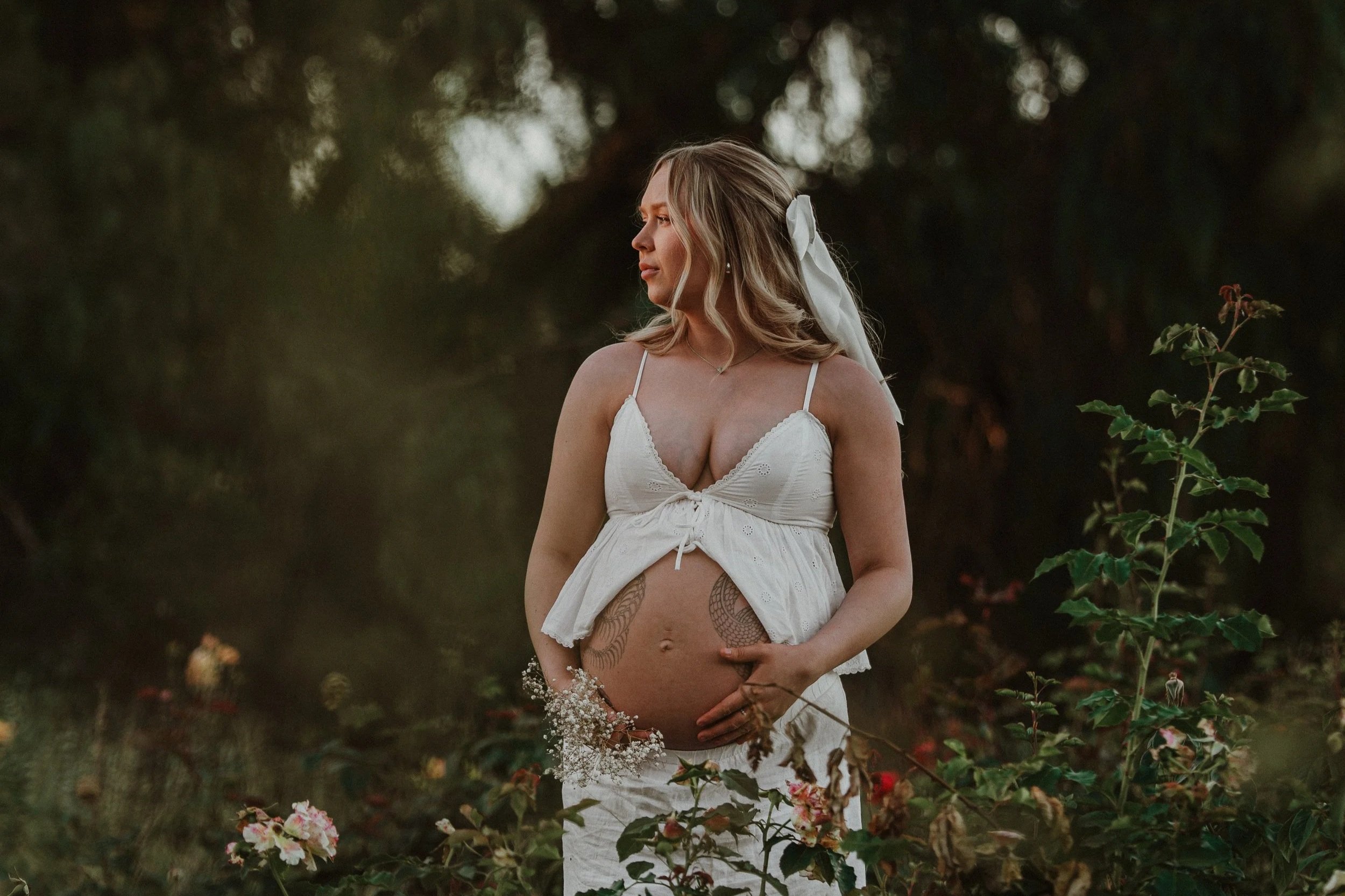 A pregnant woman wears a white dress with spaghetti straps and stands outdoors at sunset among flowering plants, with her hand on her belly, looking to the side.
