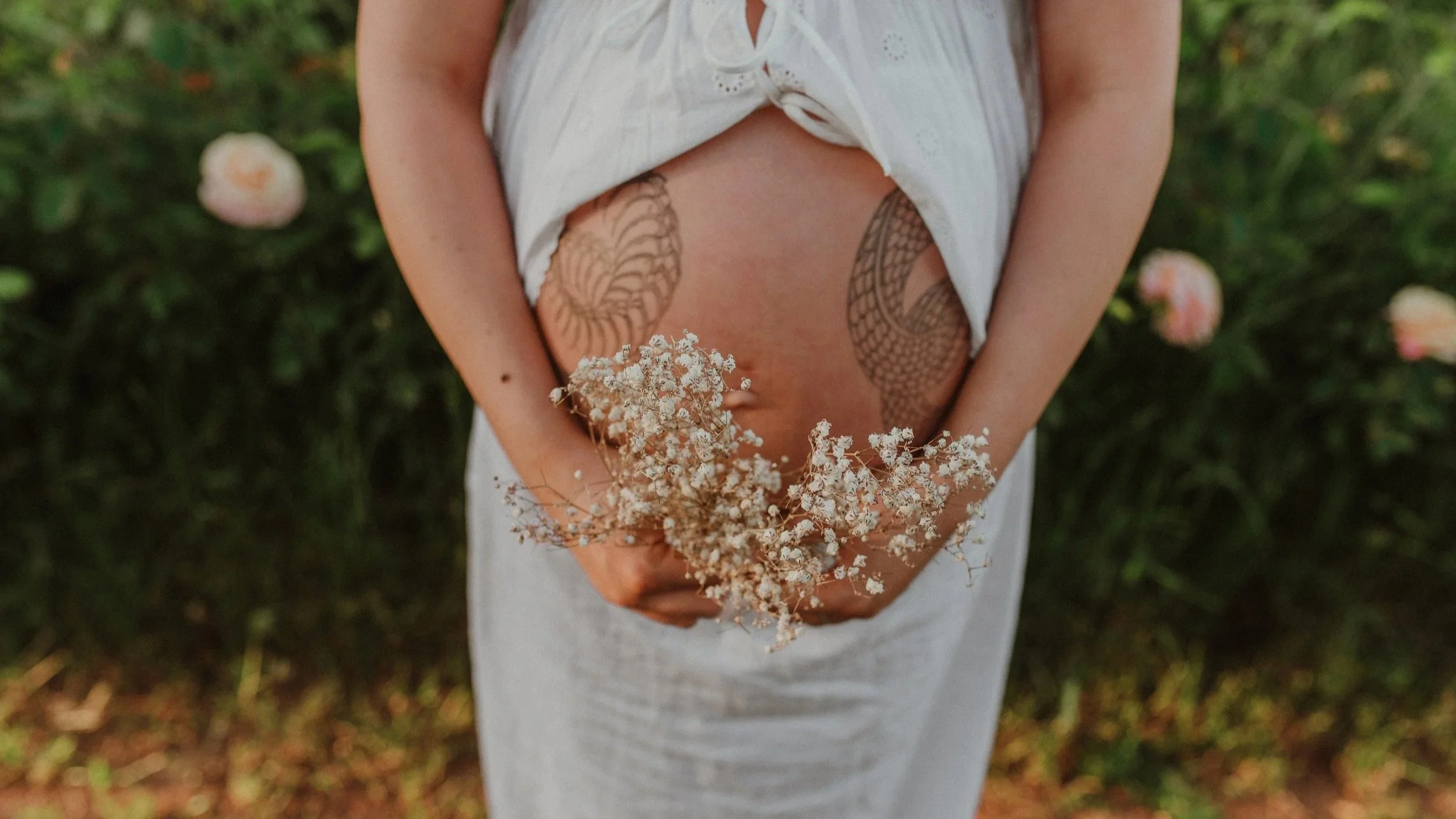 Person wearing a white dress holding a bunch of white flowers with large butterfly and snake tattoos on their stomach and sides, standing outdoors with blurred greenery background.