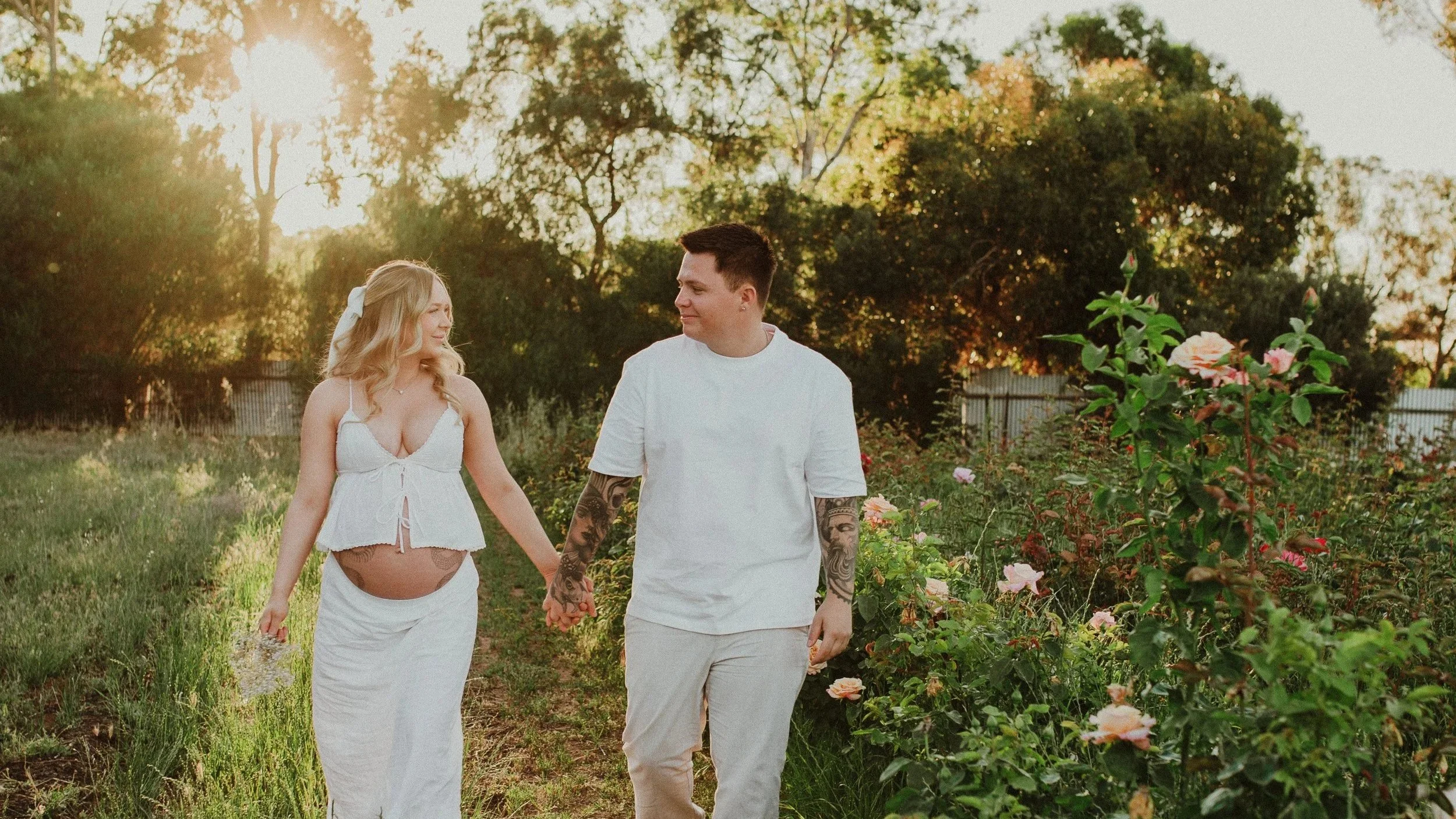 A couple walking hand in hand through a garden with blooming roses, sunlight filtering through trees in the background.