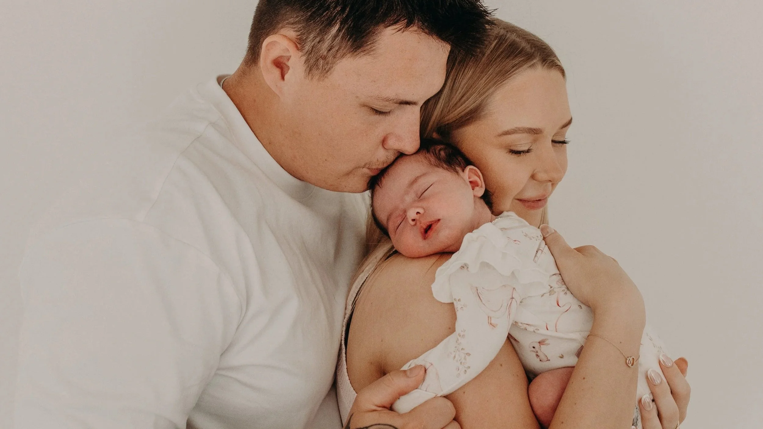 A family of three, including a man, a woman, and a newborn baby, holding the baby close as everyone gently rests their heads together with eyes closed, expressing love and tenderness.