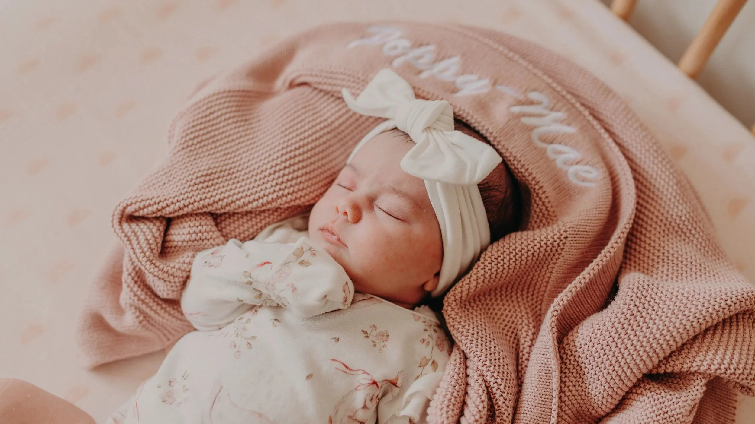 A sleeping baby wrapped in a pink knitted blanket with a white headband, lying on a soft, pink polka-dotted surface.