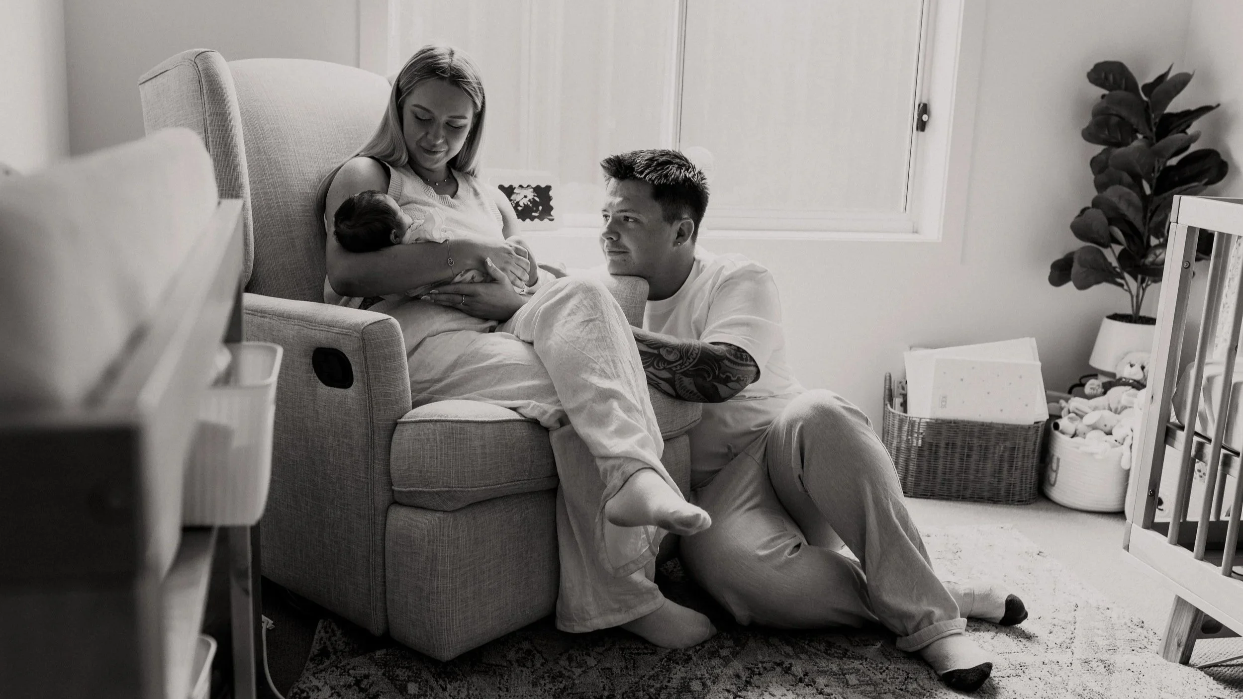 A woman sitting on a couch breastfeeding a baby, with a man kneeling beside her and looking at the baby, inside a cozy living room with a plant, window, and toys.