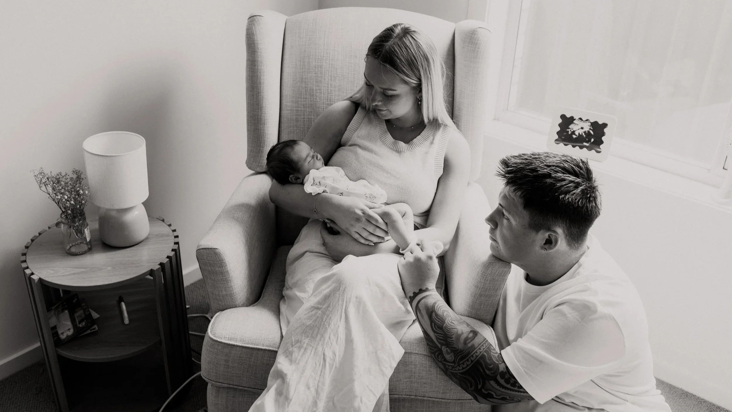 A woman holding a newborn baby while a man with tattoos assists her in a living room, with a side table of a lamp and flowers nearby.