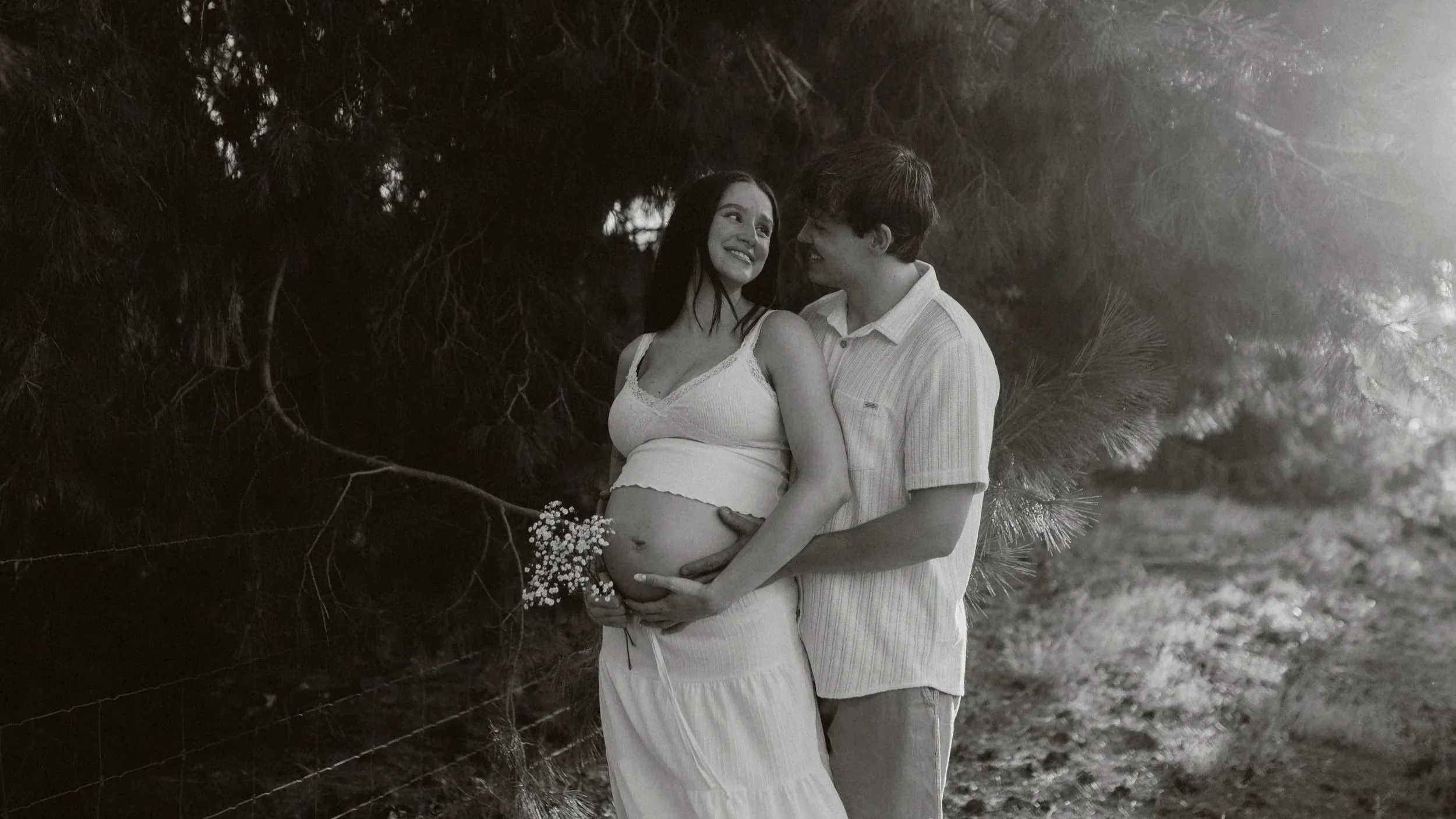 A couple standing close together outdoors, with the woman showing a pregnant belly, surrounded by trees and foliage.