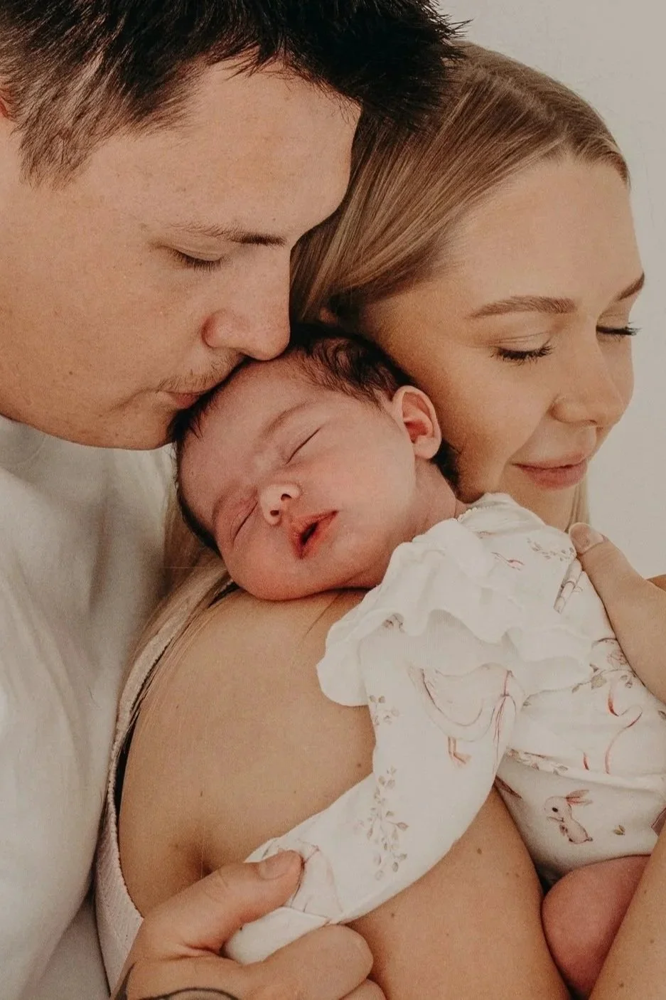 A family of three, including a man, a woman, and a newborn baby, holding the baby close as everyone gently rests their heads together with eyes closed, expressing love and tenderness.