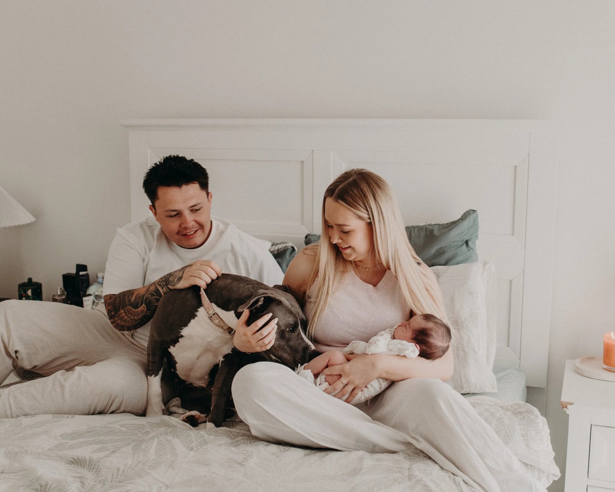 A family of four with a dog and a baby sitting on a bed in a bright bedroom, smiling and engaging with each other.