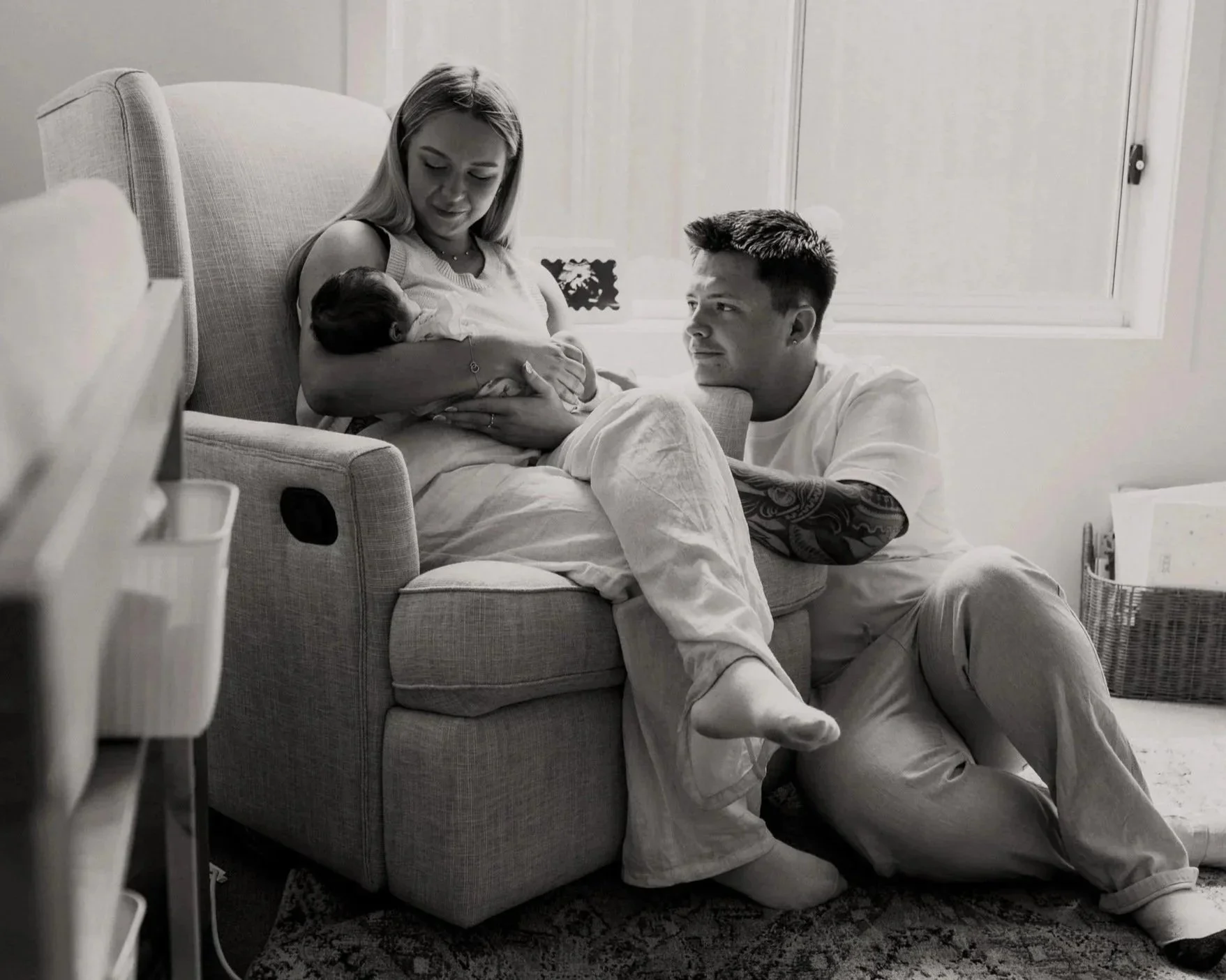 A woman sitting on a couch breastfeeding a baby, with a man kneeling beside her and looking at the baby, inside a cozy living room with a plant, window, and toys.