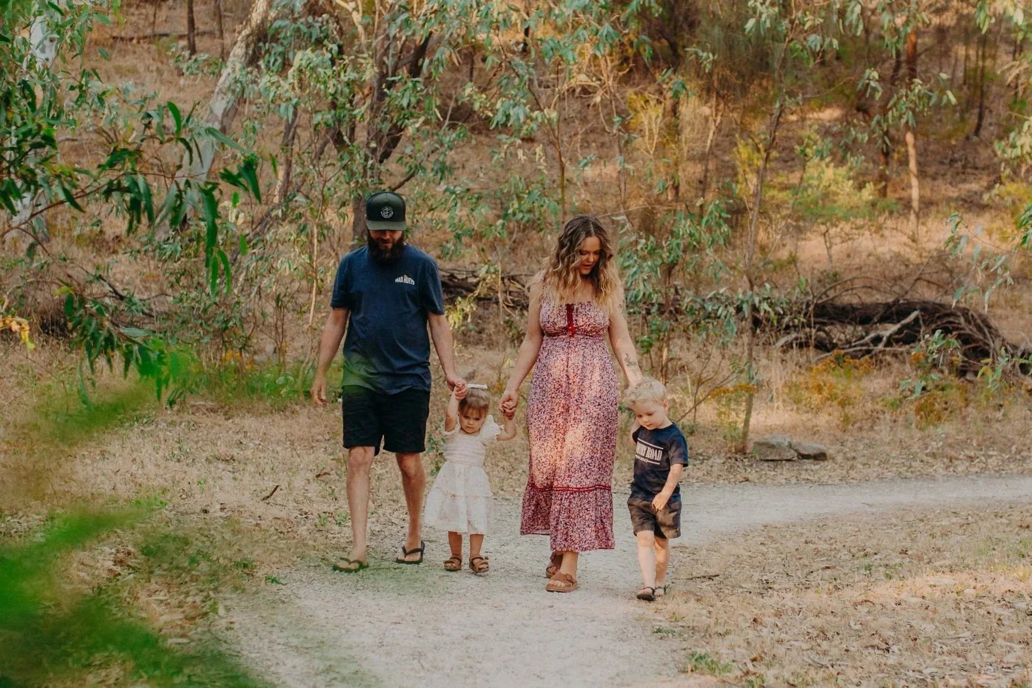 A family of four walking on a dirt path in a wooded area. The father is wearing a dark blue t-shirt and shorts. The mother is wearing a long floral dress. Two young children, a girl in a white dress and a boy in a dark T-shirt, are holding their hand