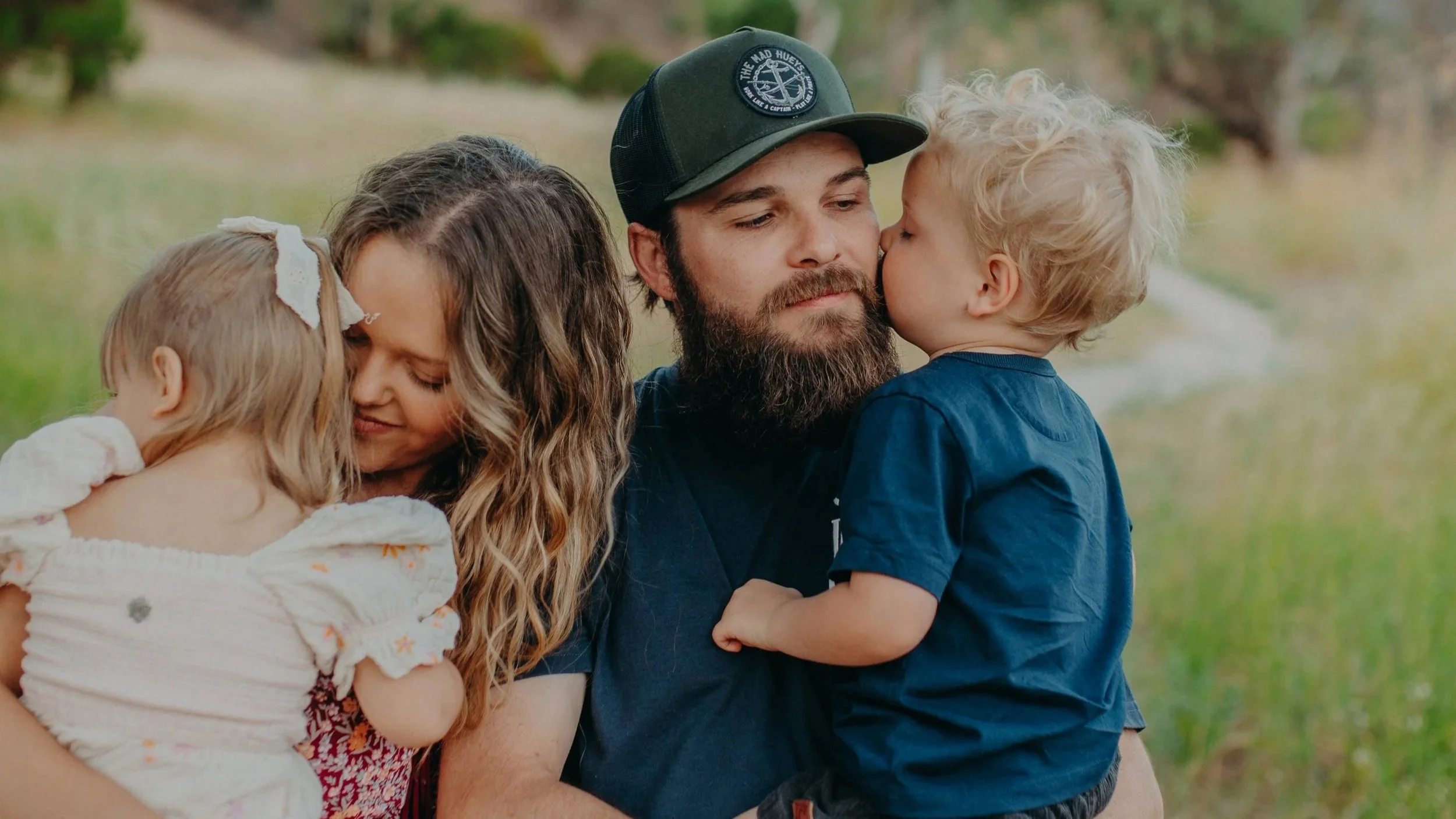 A man with a beard wearing a black baseball cap hugging a young girl with curly blonde hair, a woman with long wavy hair holding a young girl with short blonde hair in a white dress, outdoors in a natural setting.