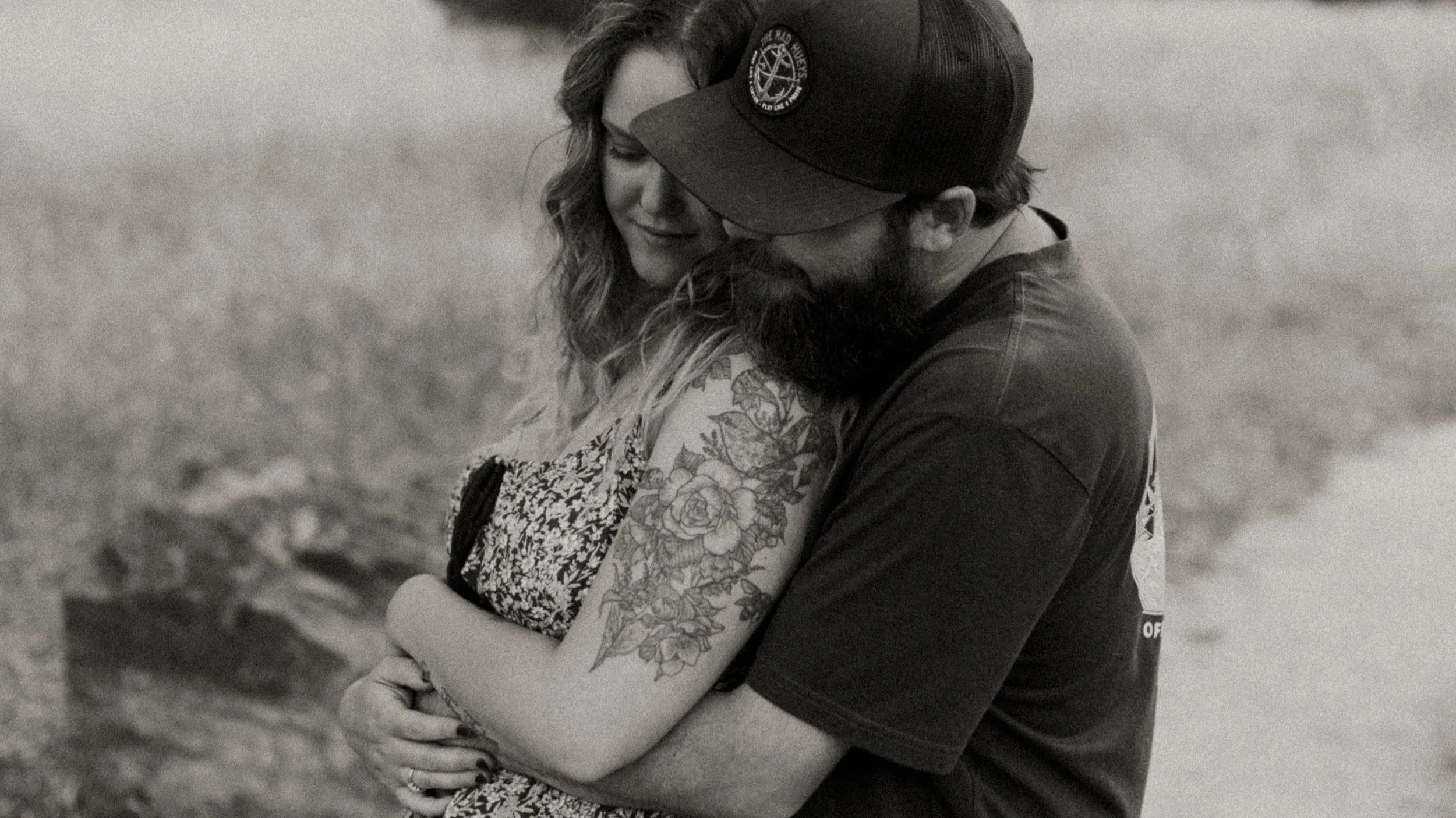 A man and woman embrace outdoors, with the woman's arm tattoo visible, and both have their eyes closed, sharing an intimate moment.