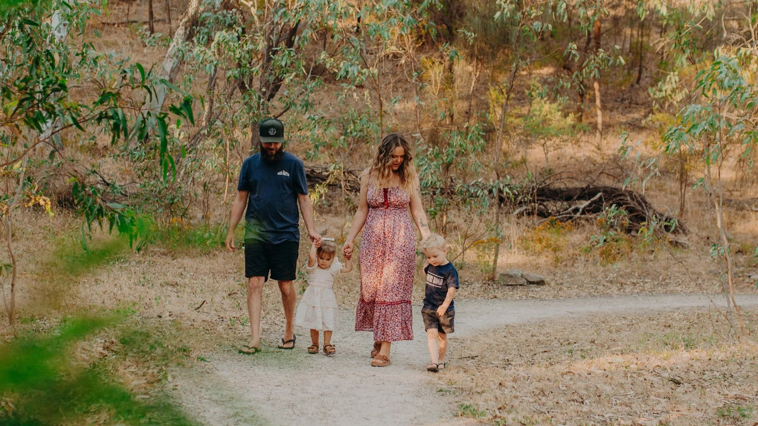 A family of four walking on a dirt path in a wooded area. The father is wearing a dark blue t-shirt and shorts. The mother is wearing a long floral dress. Two young children, a girl in a white dress and a boy in a dark T-shirt, are holding their hand