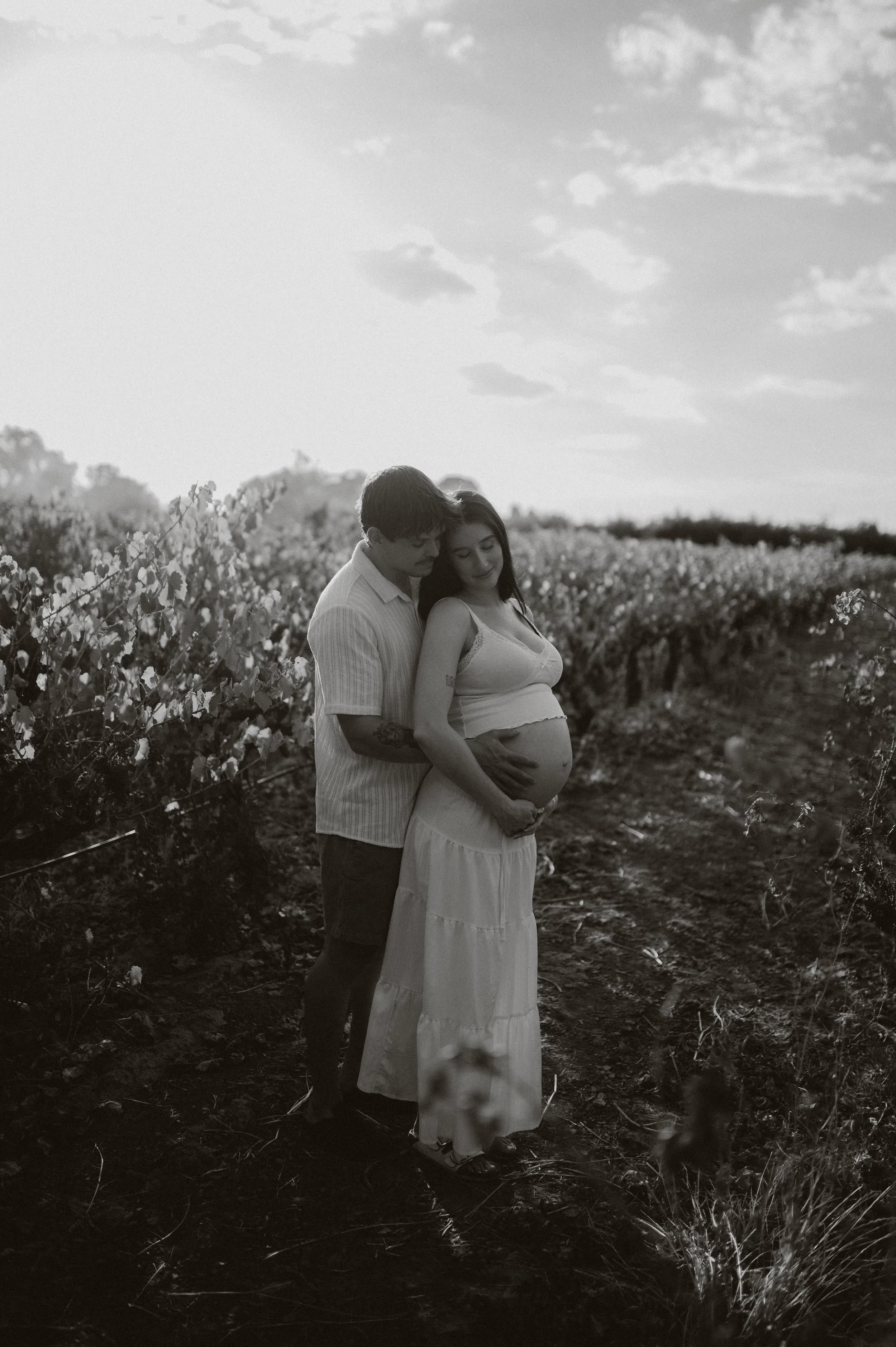 A pregnant woman and a man standing close together in a field of flowers during sunset, with the man gently resting his hands on her belly and both looking down at it.