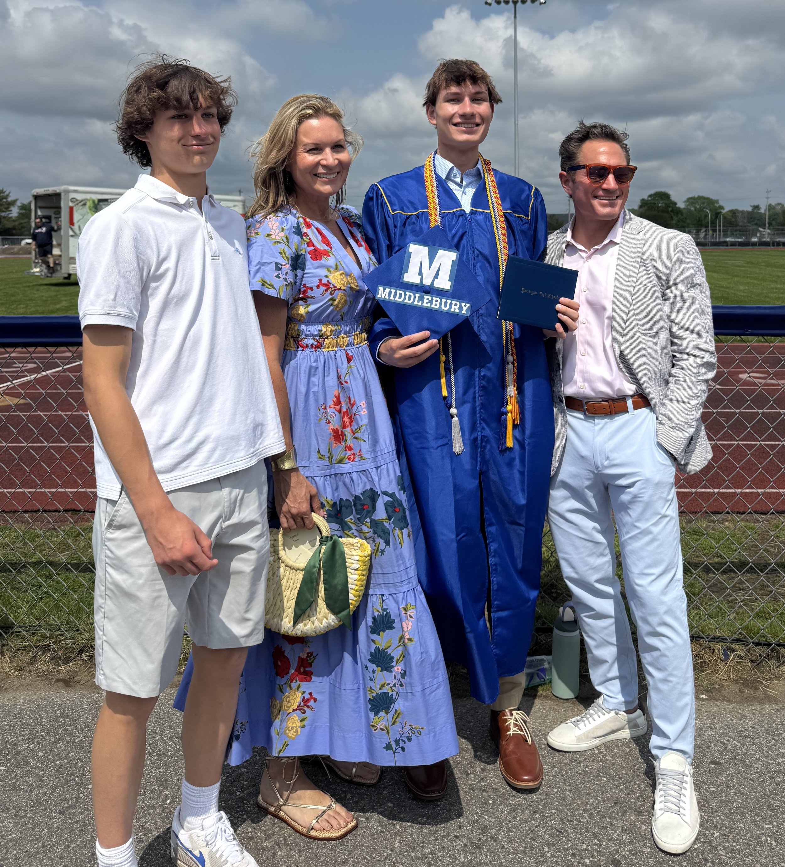 Four people standing in a row outdoors, during a graduation ceremony. The graduate is dressed in a blue cap and gown with medals, holding a diploma. The others are dressed in casual and semi-formal attire, standing next to the graduate. The background shows a sports field and cloudy sky.