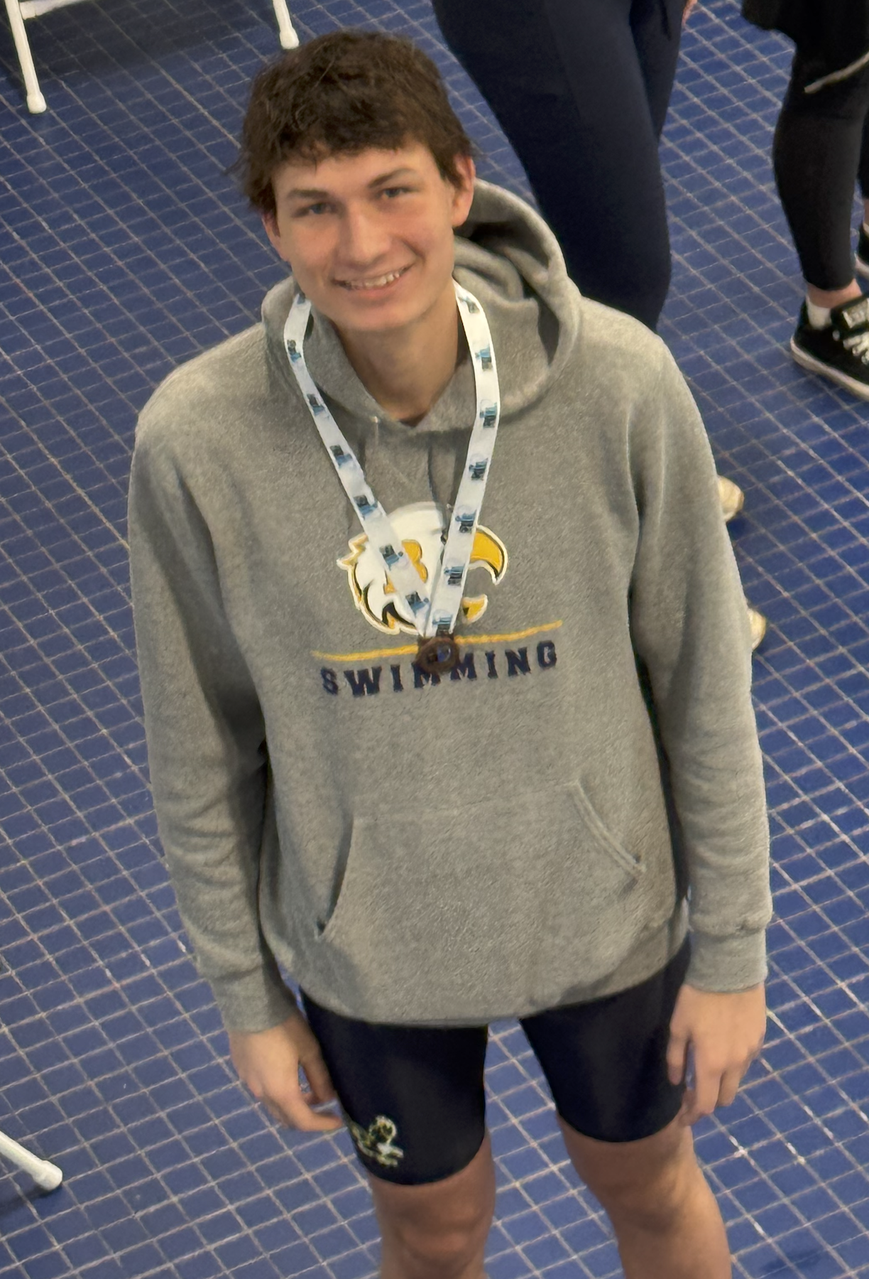 Young man standing indoors on a blue patterned carpet, wearing a gray hoodie with a swimming logo, a lanyard around his neck, and black shorts with a small logo, smiling at the camera.