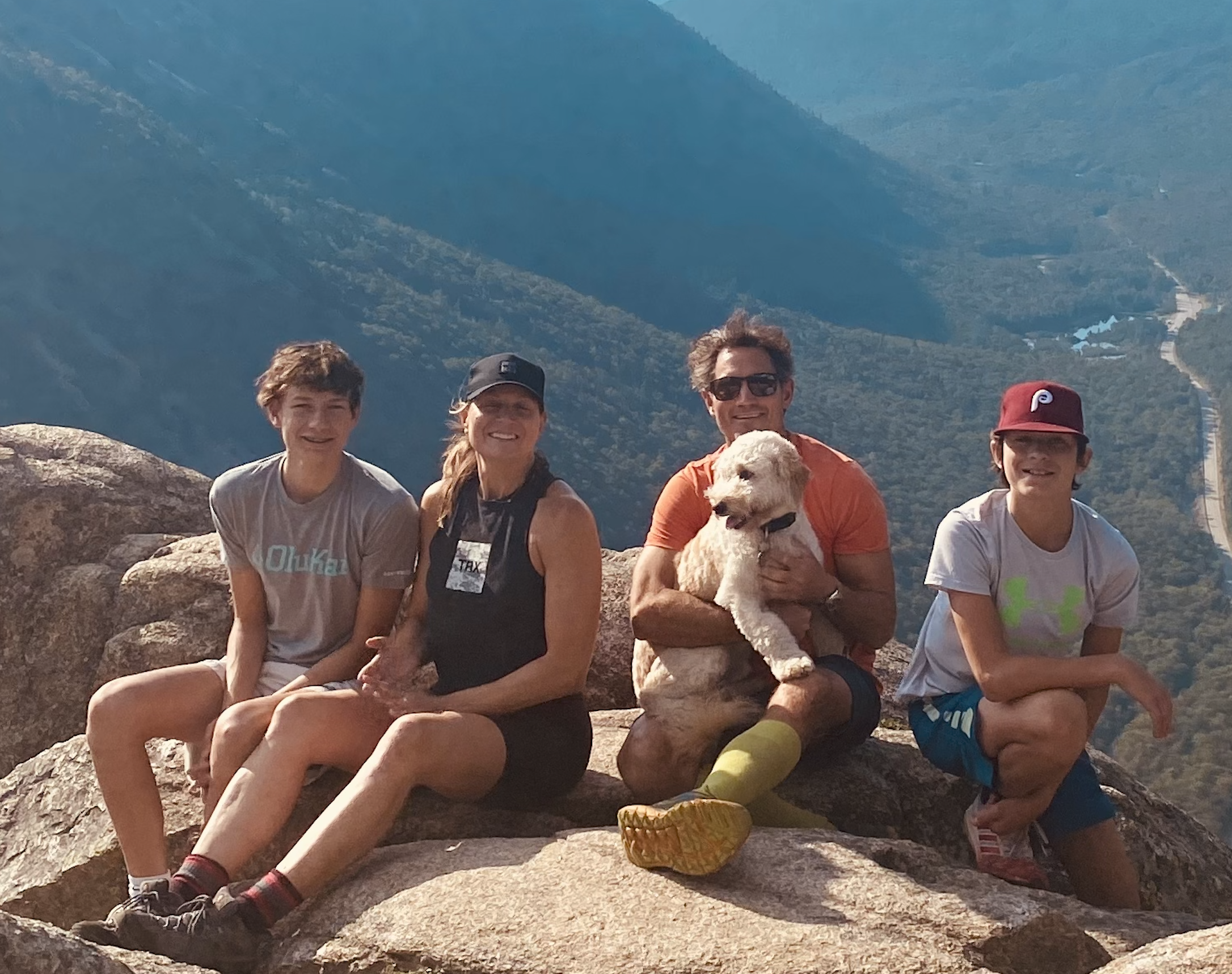 Four people and a dog sitting on a large rock at a scenic overlook with mountains and winding road in the background.