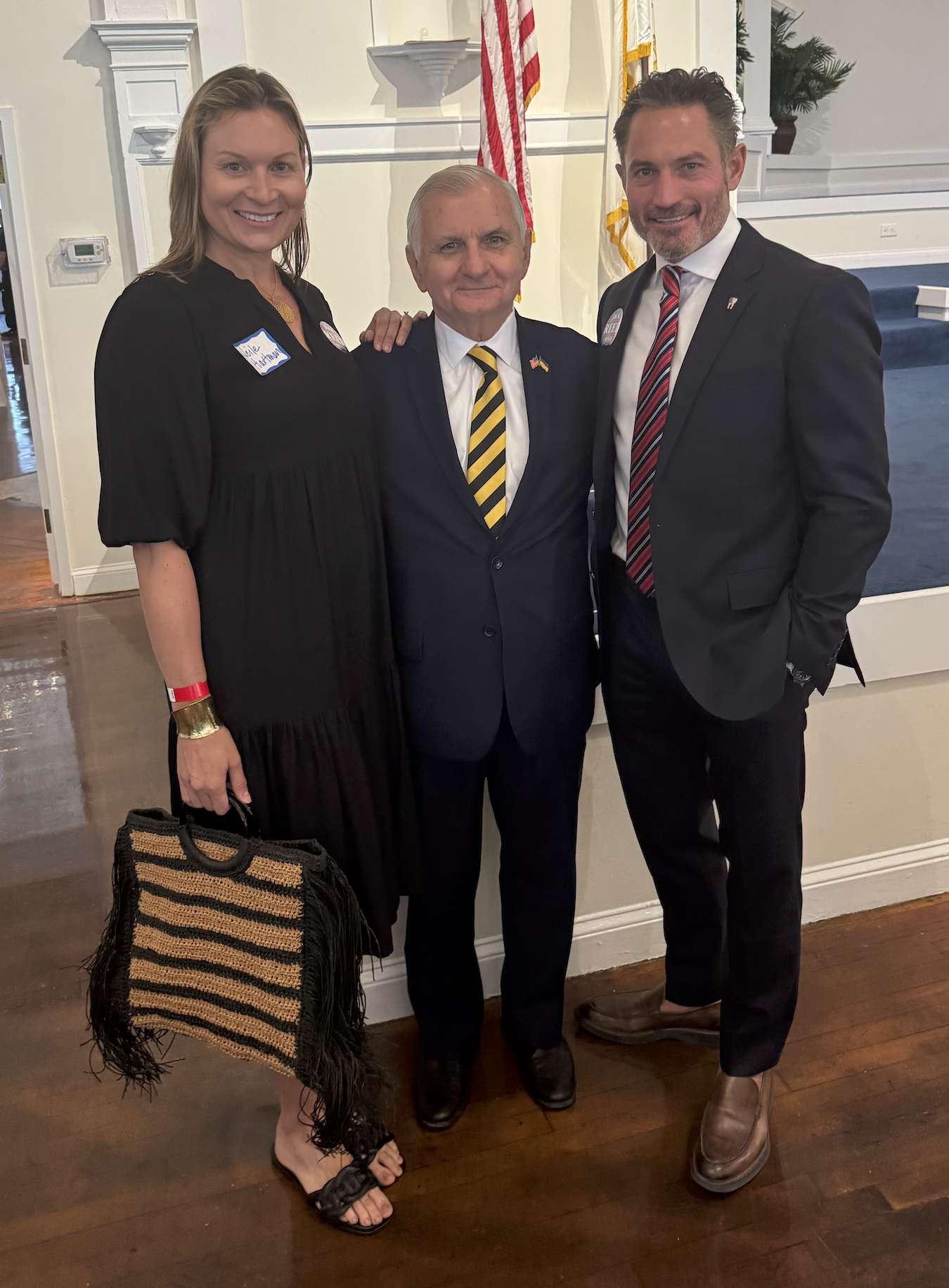Three people standing together in a room with white walls and American flags in the background, smiling at the camera. They are Neile Hartman (left), U.S. Senator Jack Reed (center) and Dr. Frederick Hartman.