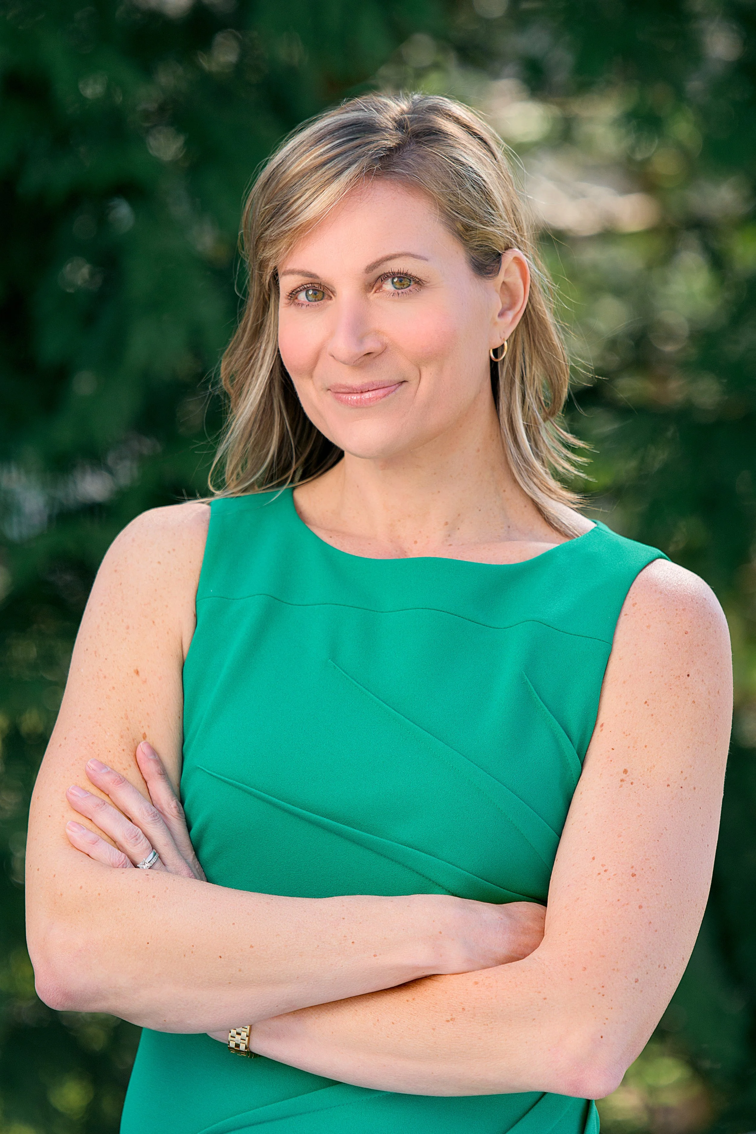 A woman with blonde hair, green eyes, and light skin standing outdoors with green blurred background, wearing a sleeveless green dress, a gold watch, a ring, and small hoop earrings, with arms crossed.