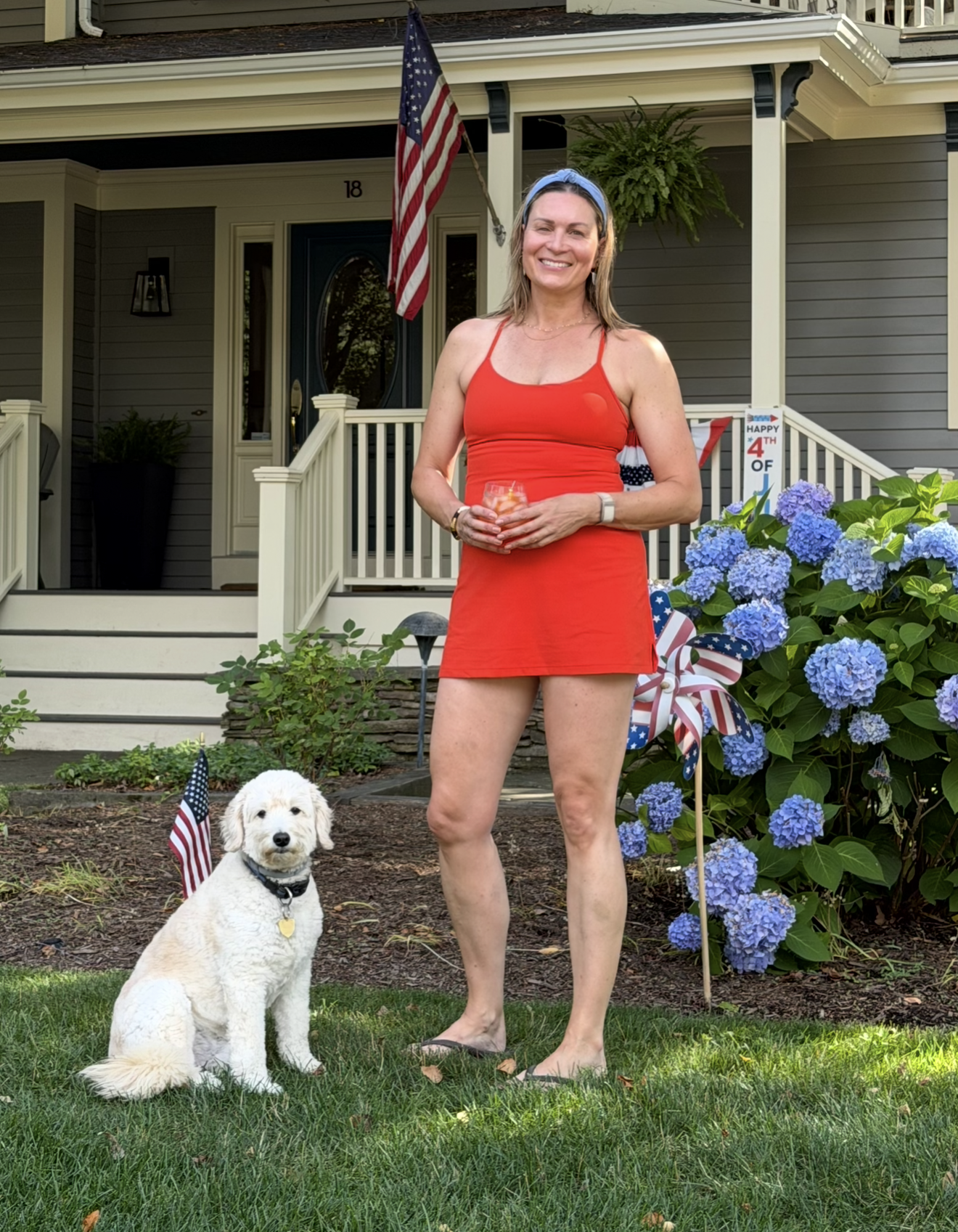 A smiling woman in a red dress and headband standing in a garden in front of a house with an American flag. A white dog sits nearby with a small American flag on the ground. The garden has blue hydrangeas and an American-themed pinwheel.