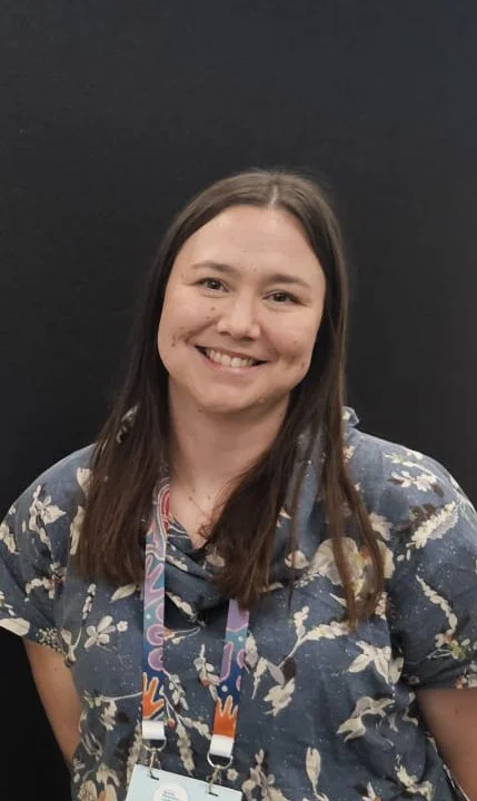 Woman with long brown hair smiling, wearing a blue floral shirt and a conference lanyard, standing against a black background.