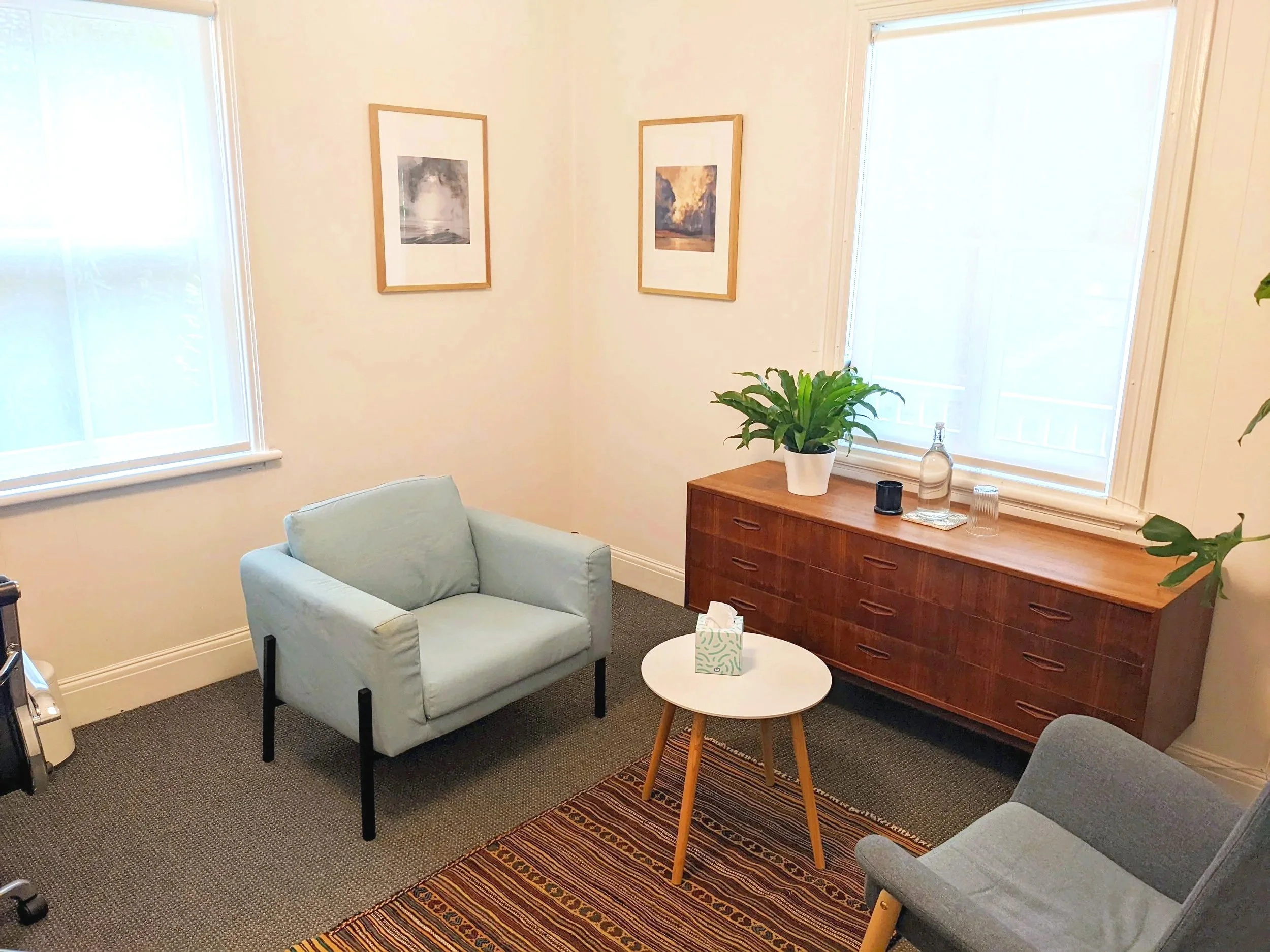 A cozy seating area with two light blue armchairs, a small round white table, a wooden sideboard with a potted plant, bottles, and glasses, sunlight coming through two large windows, framed artwork on the walls, and a patterned rug on the carpeted floor.