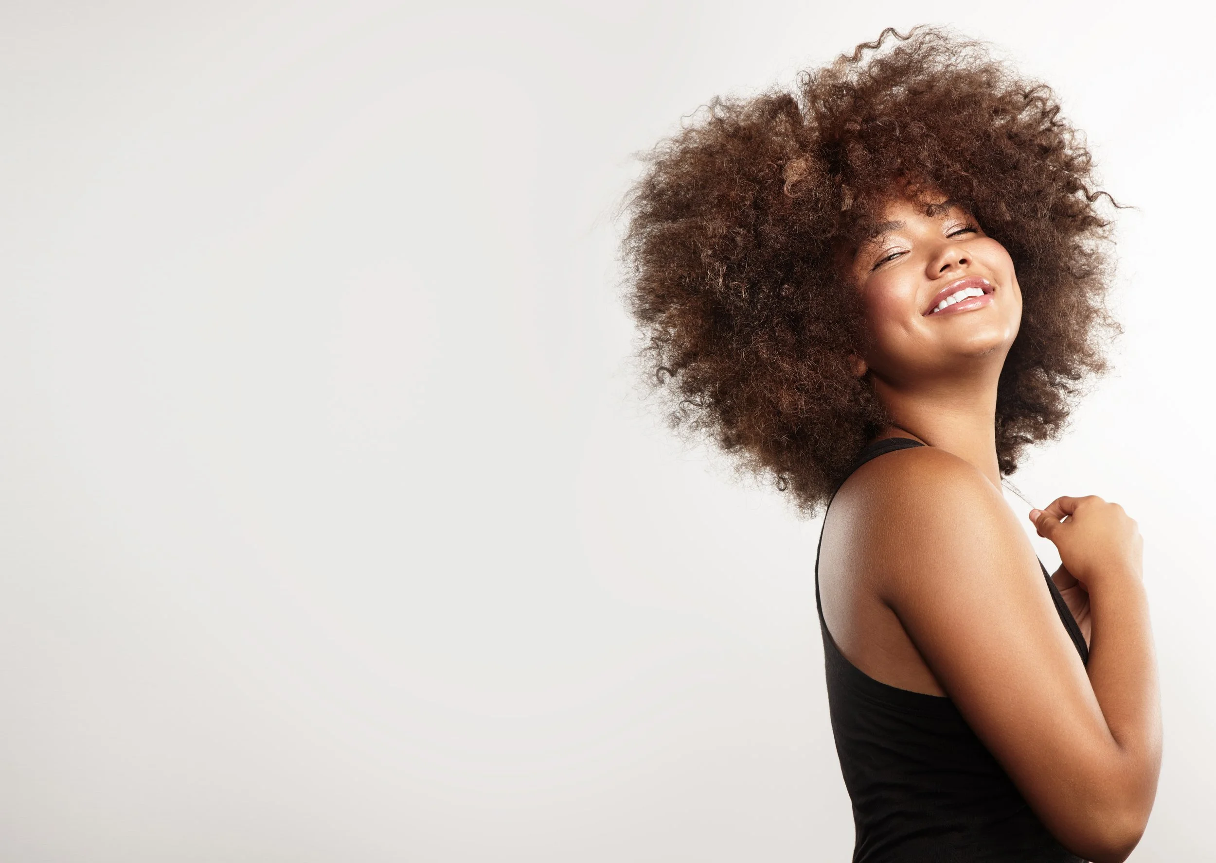 A woman with a large, curly afro hairstyle, wearing a black tank top, smiling with closed eyes, standing against a plain white background.