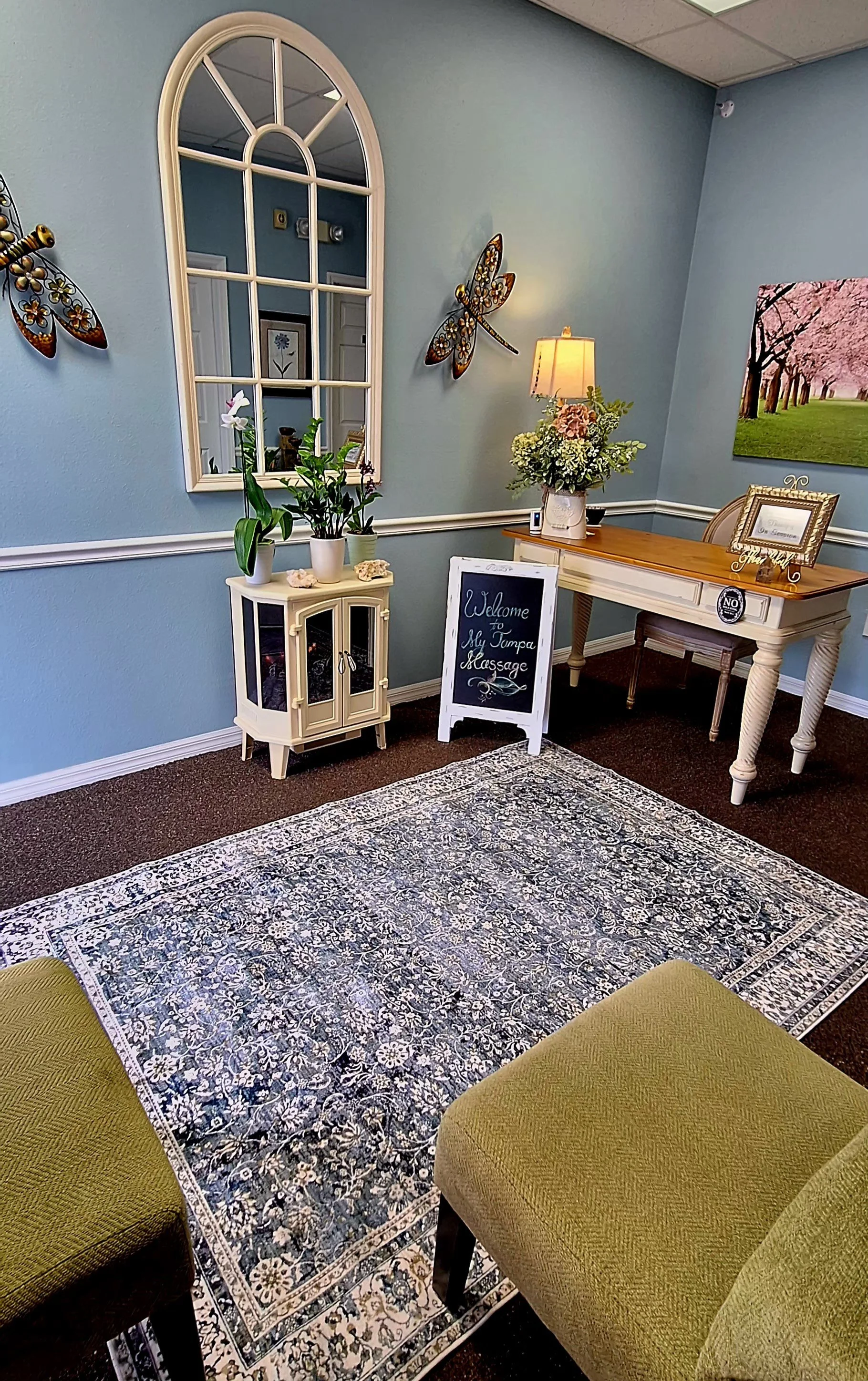 Inside a waiting room, there are two green tufted chairs, a black and white patterned rug, a white cabinet with plants, a window with a decorative mirror, a card table with a floral arrangement, a picture frame, and wall decorations including a sign 