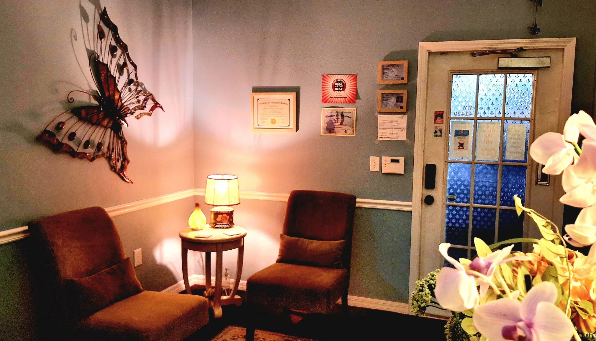 A cozy living room corner with two brown upholstered chairs, a small round wooden table with a lamp, a framed certificate on the wall, several smaller framed pictures, a doorway with glass panes, and a large decorative butterfly wall art. There are f