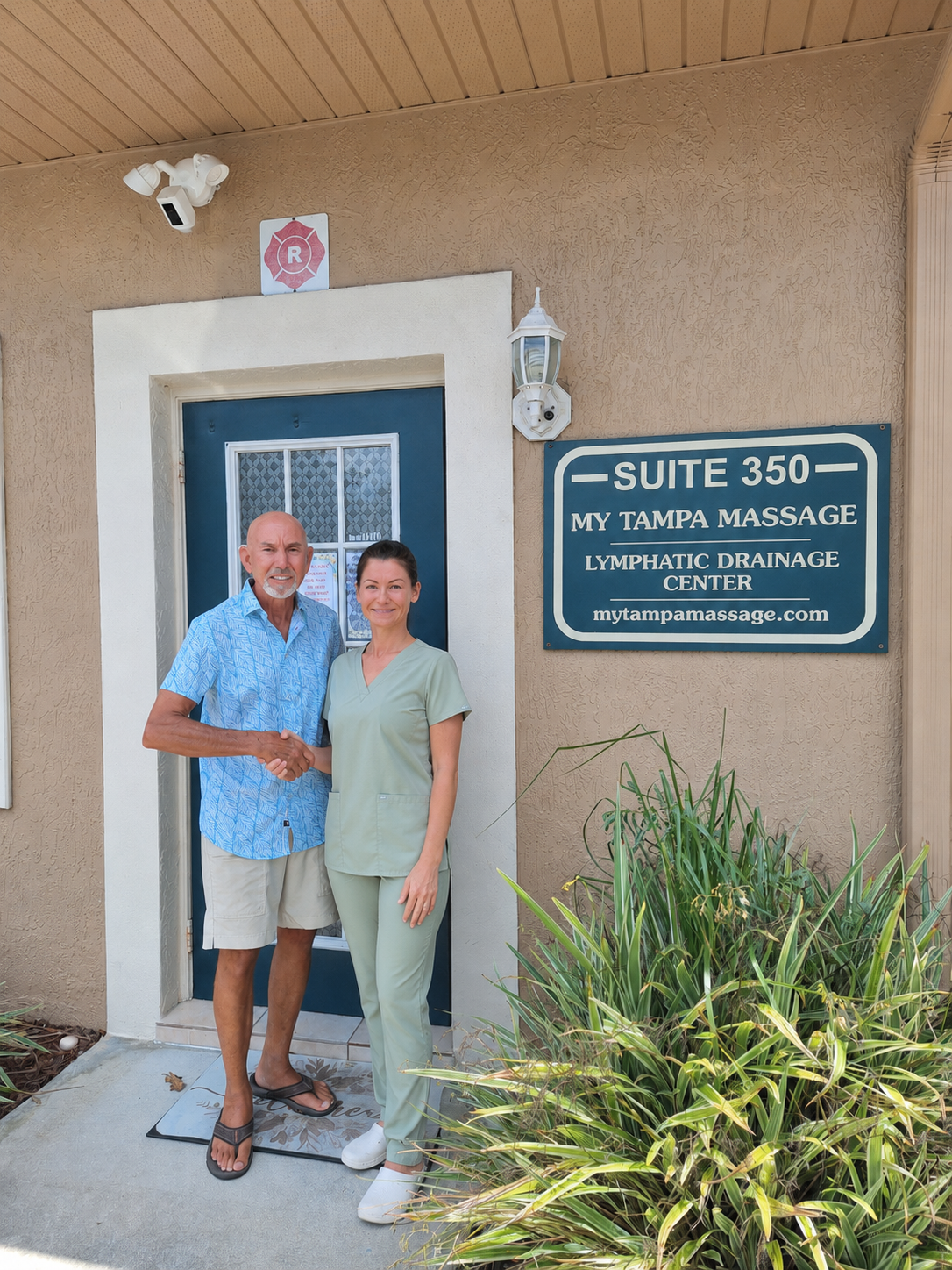 A man and woman shaking hands in front of a door outside a massage therapy clinic. The man is wearing a blue patterned shirt and beige shorts with flip-flops. The woman is dressed in light green scrubs and white shoes. A sign next to them reads 'Suite 350 My Tampa Massage Lymphatic Drainage Center' with a website address.