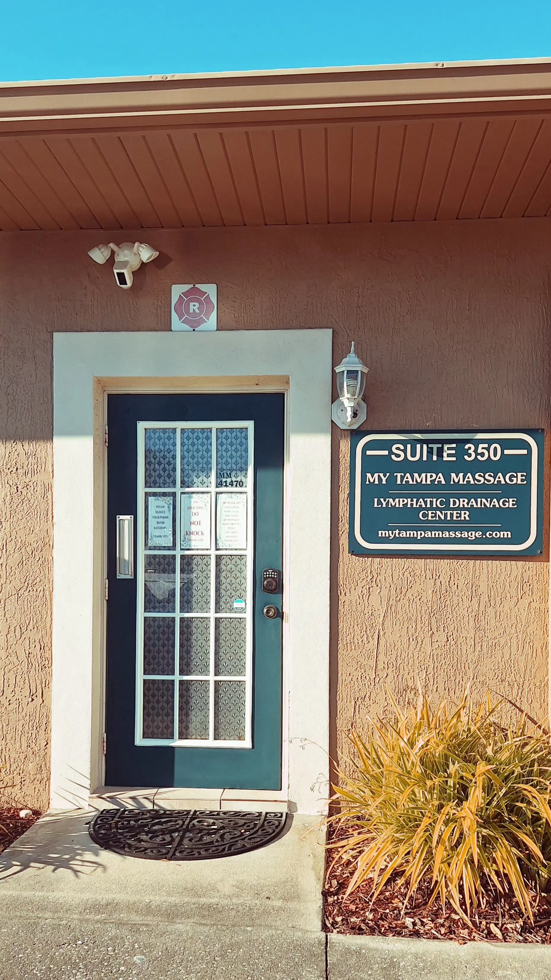 Office entrance with a blue door, white frame, security keypad, and a sign that reads 'Suite 350, My Tampa Massage, Lymphatic Drainage Center, mytampamassage.com'. There is an outdoor lantern and a plant with yellowish leaves to the right.