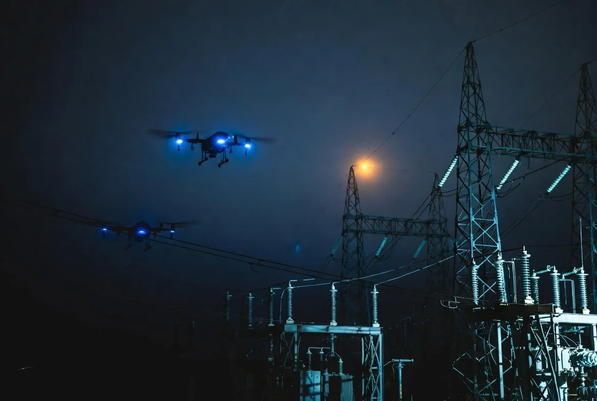 Nighttime scene showing electrical power lines and towers with two drones flying in the dark sky, illuminated by blue and yellow lights.
