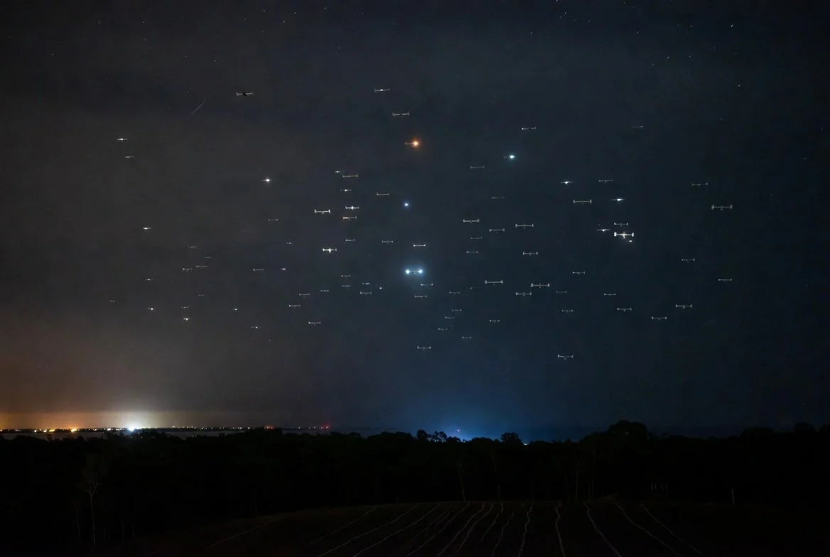 Night sky filled with many B-52 aircraft flying in formation, with some bright lights and distant city lights near the horizon.