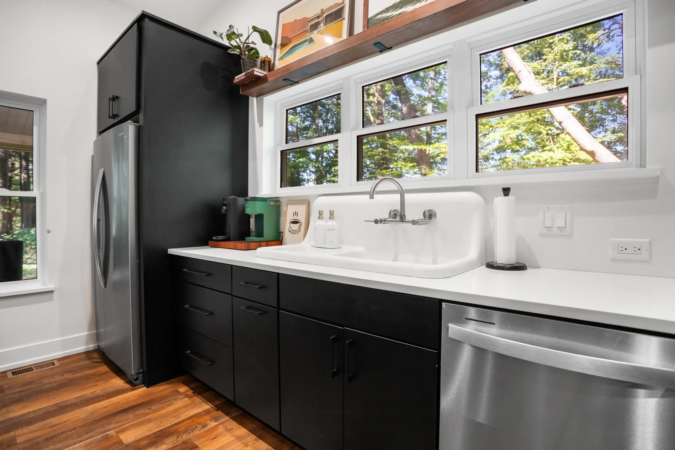 Modern kitchen with black cabinets, white countertop, stainless steel fridge, white farmhouse-style sink, and large window with a view of trees.