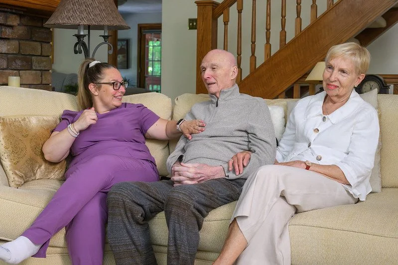 Three people sitting on a cream-colored couch in a living room, engaging in conversation. A woman caregiver in purple scrubs with glasses, an elderly man in a gray zip-up sweater, and an elderly woman in a white blouse and pants.