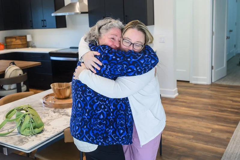 Two women hugging warmly in a modern kitchen, one older with gray hair and the other younger with glasses, both smiling with joy.