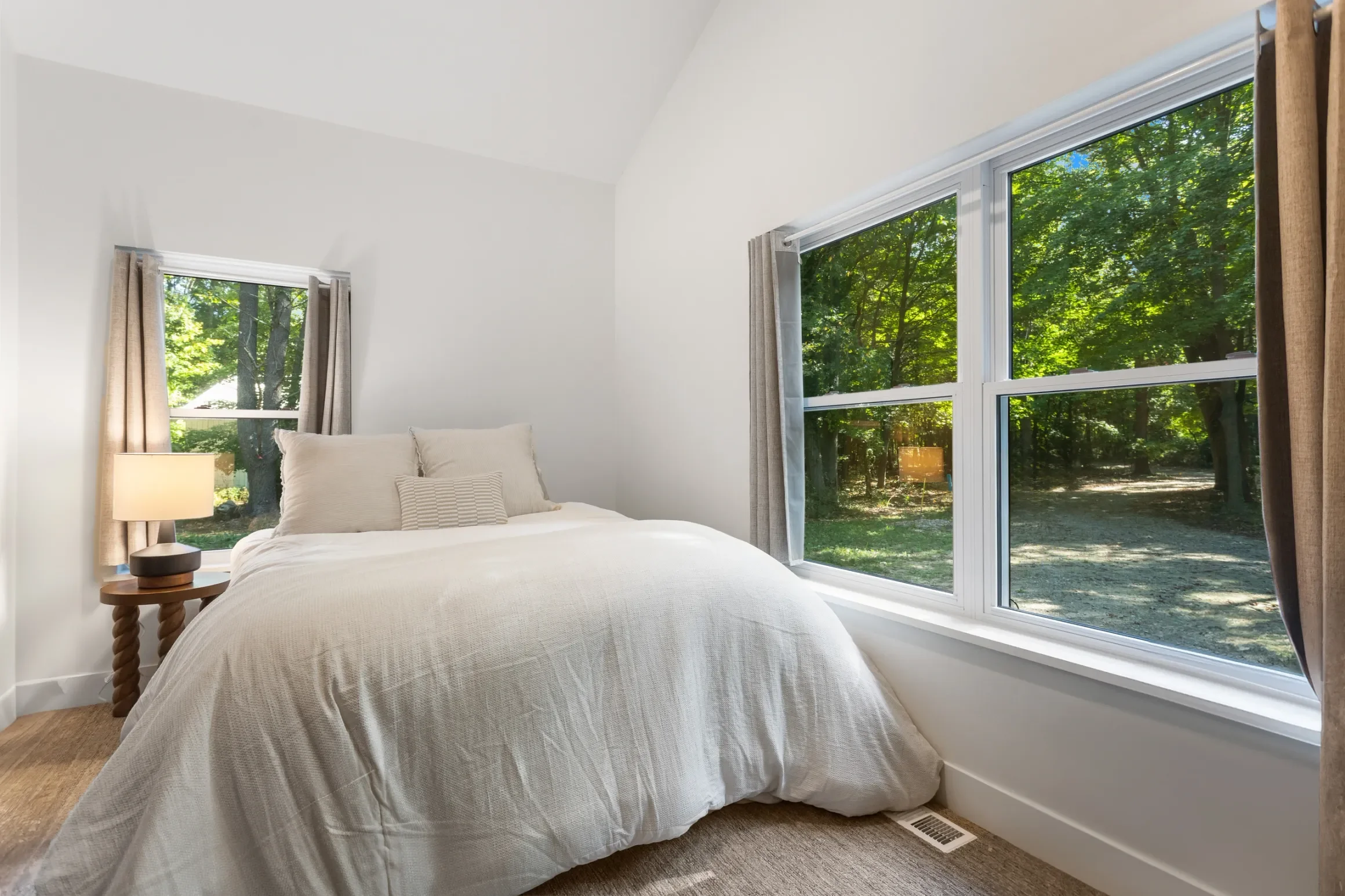 A bedroom with a large window showing green trees outside, a bed with white bedding and pillows, a bedside table with a lamp, and beige curtains.