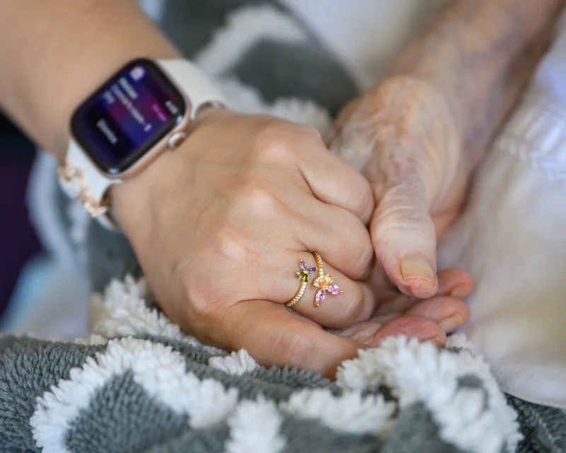 Close-up of two elderly hands clasped, one wearing a smartwatch and colorful butterfly-shaped rings, resting on a soft, fluffy blanket.