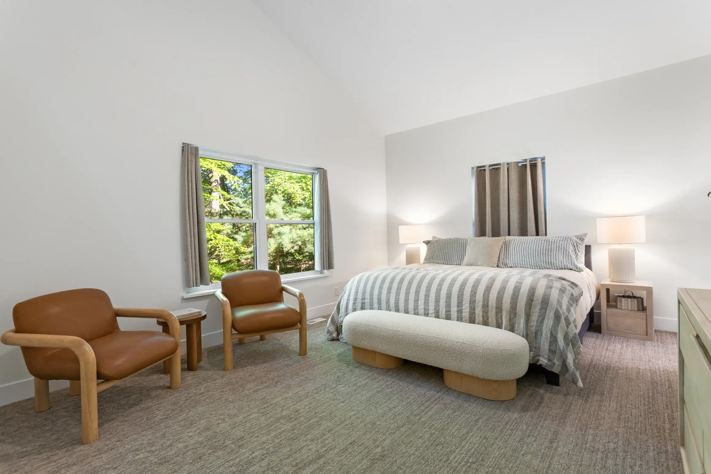 Clean bedroom with large window showing greenery outside, king bed with striped bedding, two bedside lamps, tan chairs, and a small white table.