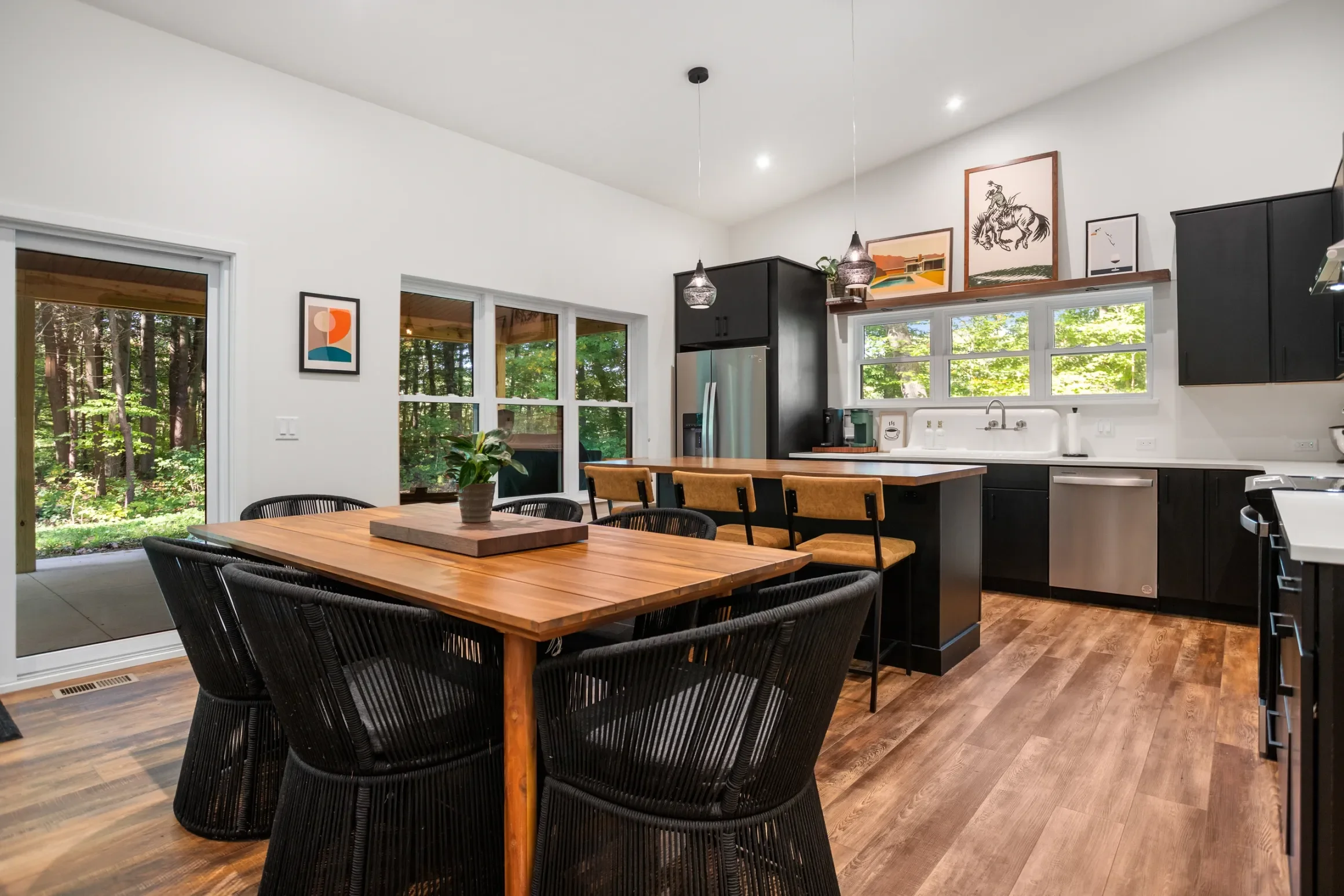 Modern kitchen with black cabinets, wooden dining table with black chairs, large windows letting in natural light, artwork on the wall, pendant lighting, and a view of a wooded outdoor area.