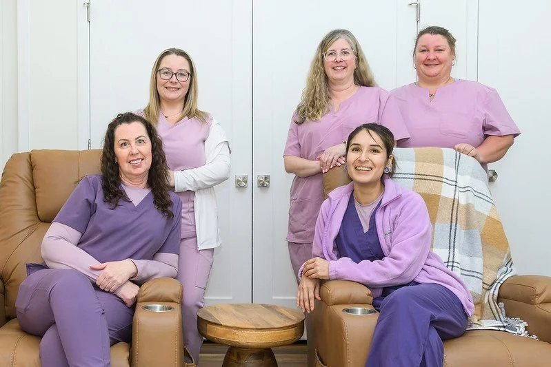 Group of five women in medical scrubs smiling in a living room, with some sitting on leather recliners and others standing behind them.