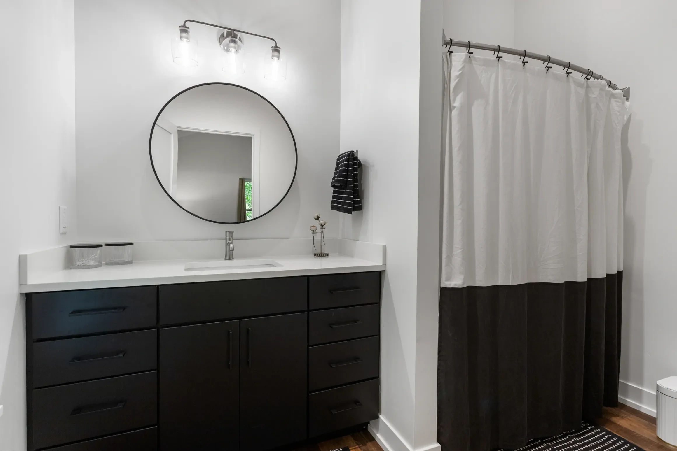 Modern bathroom with a white vanity, black cabinets, round mirror, a shower, and white shower curtain, with medium-tone wood flooring.