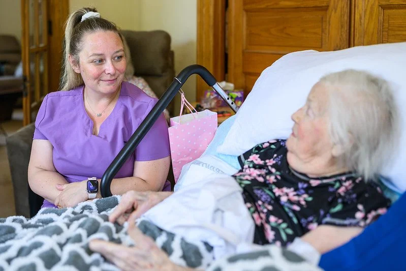 A caregiver sitting beside an elderly woman in a hospital bed, looking at each other and smiling.