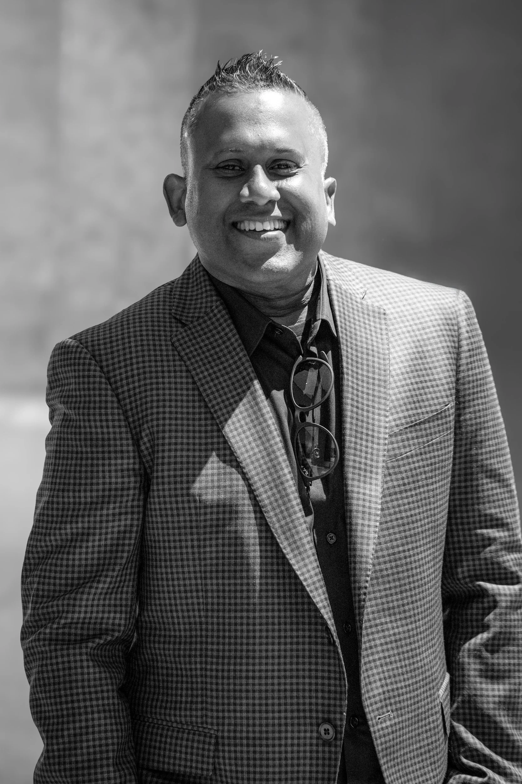 A smiling man wearing a checkered blazer and dark shirt, with sunglasses hanging from his collar, standing outdoors against a blurred background.