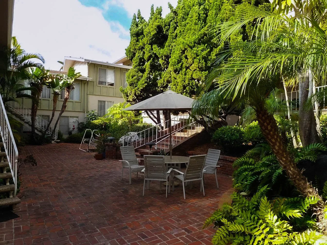 A brick patio in a lush garden with white outdoor furniture and a large white umbrella, surrounded by green trees and plants.